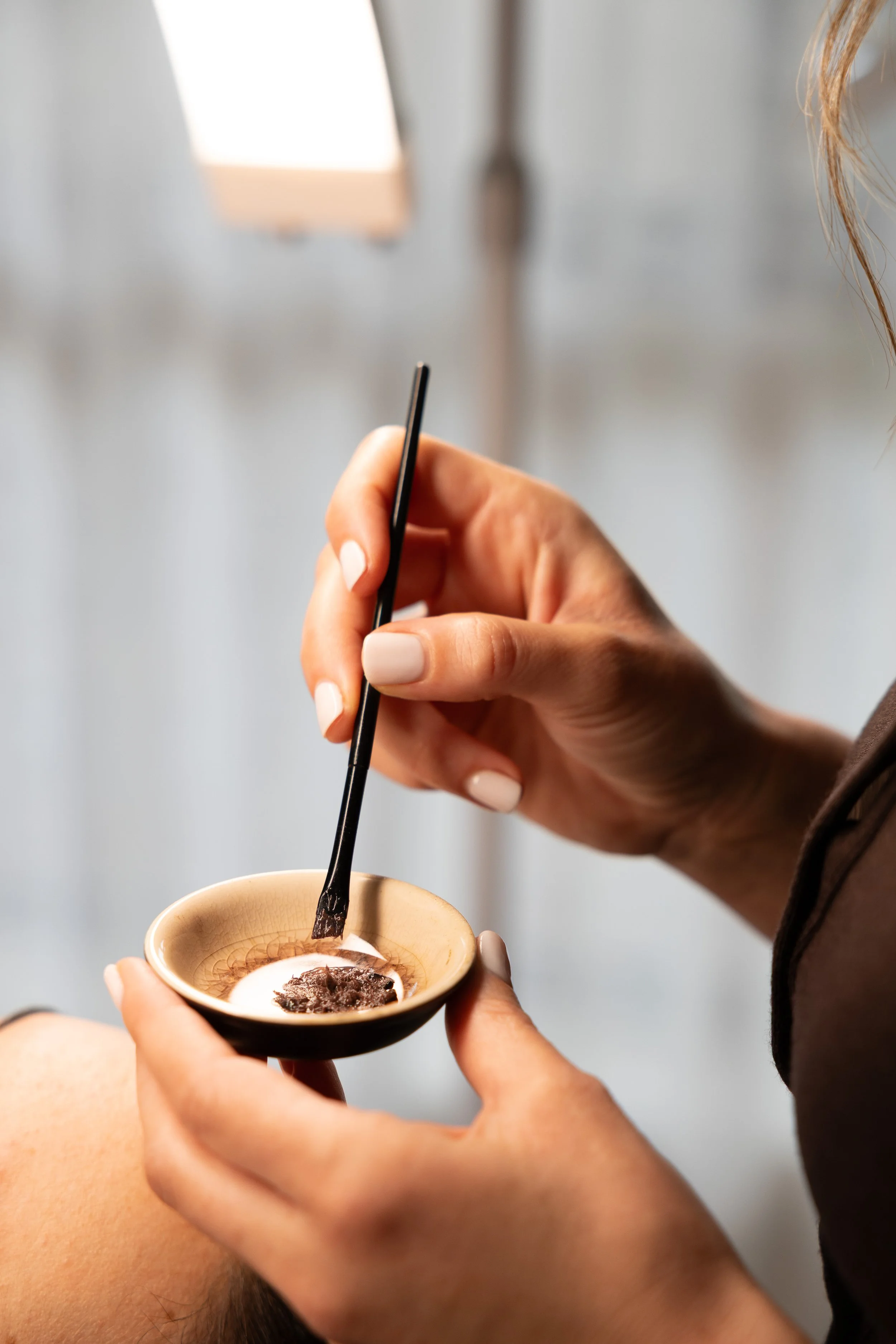 A person applying a face mask or skincare treatment to another person's face using a small brush, with a small bowl holding the skincare product.