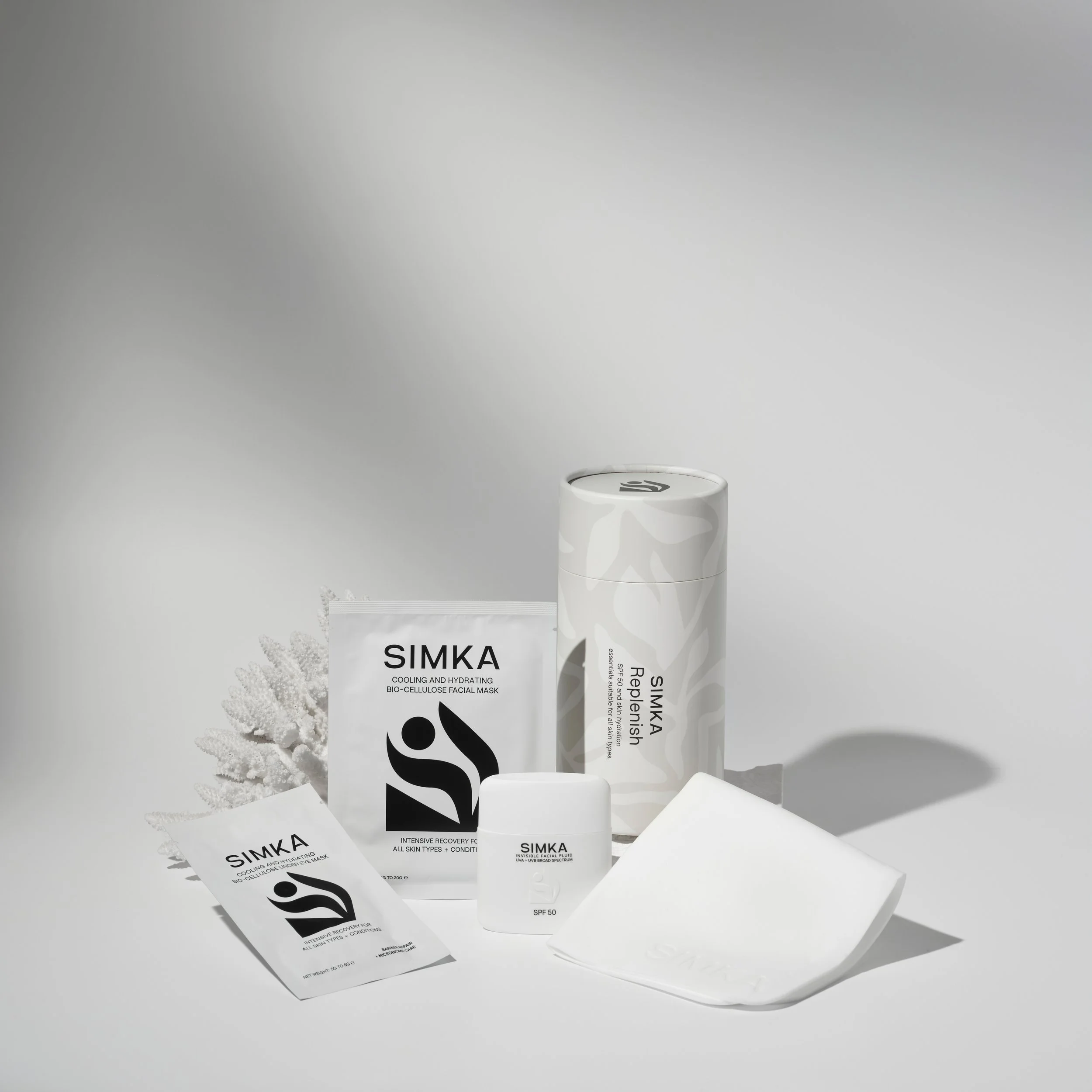 Assorted skincare products and facial masks on a white surface, including a white bottle, a packet labeled 'SIMKA', and a tube labeled 'Replenish SPF 50' with decorative white coral.