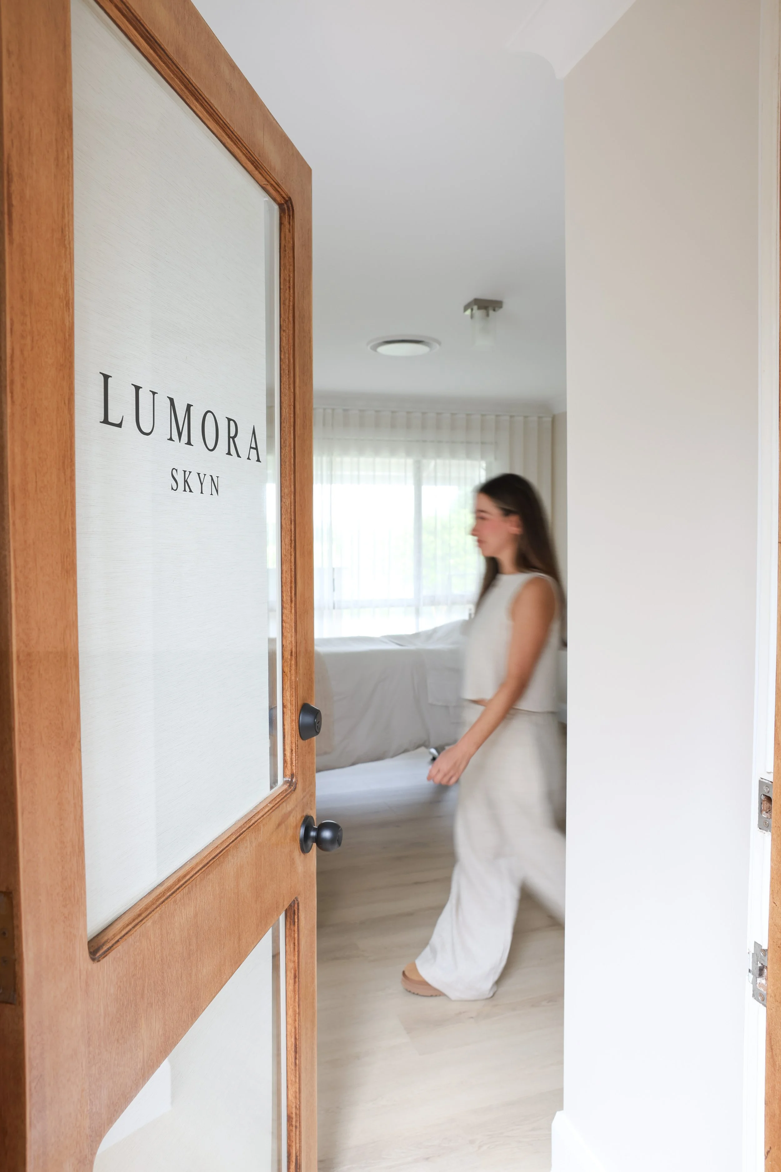 A woman in a white dress entering a bedroom, seen through an open wooden door with a frosted glass panel labeled "LUMORA SKYN." The bedroom has light-colored walls, sheer curtains, and a bed with white bedding.