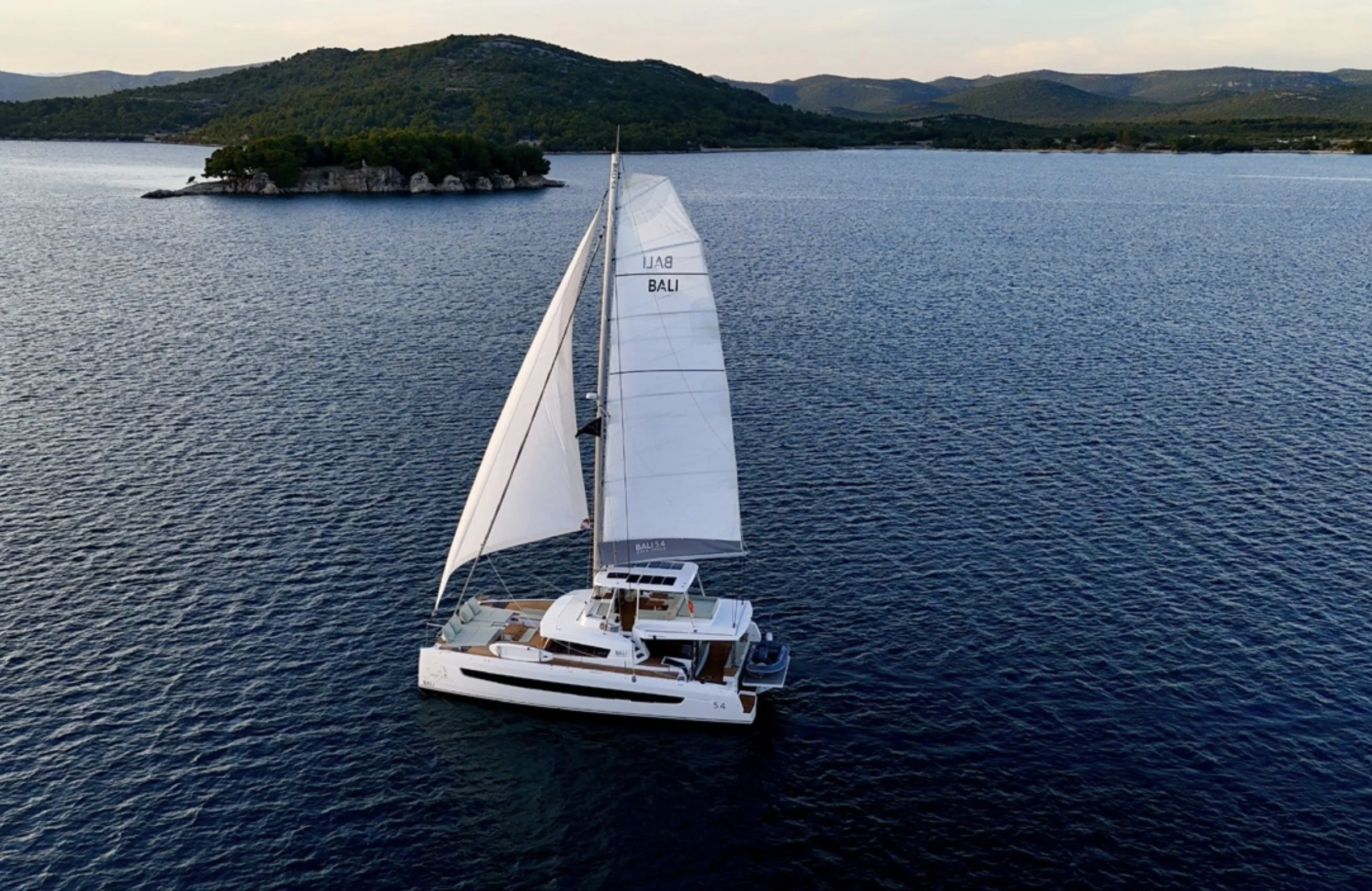 A white sailing yacht with large sails on a large body of water, with tree-covered islands and mountains in the background.