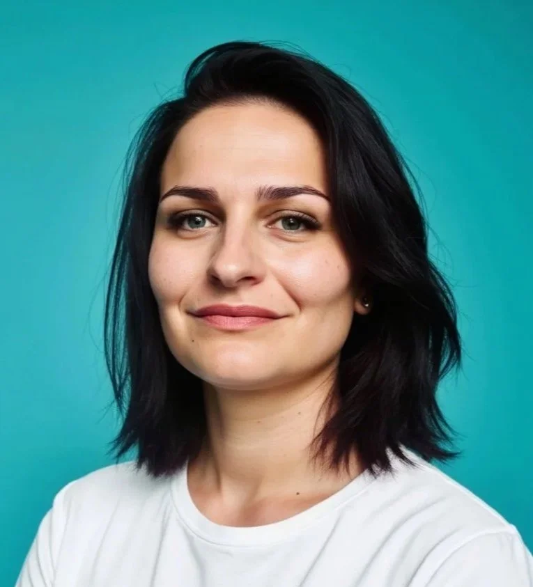 Eine junge Frau mit schwarzen, schulterlangen Haaren und einem weißen T-Shirt steht vor einem blauen Hintergrund, lächelt leicht.