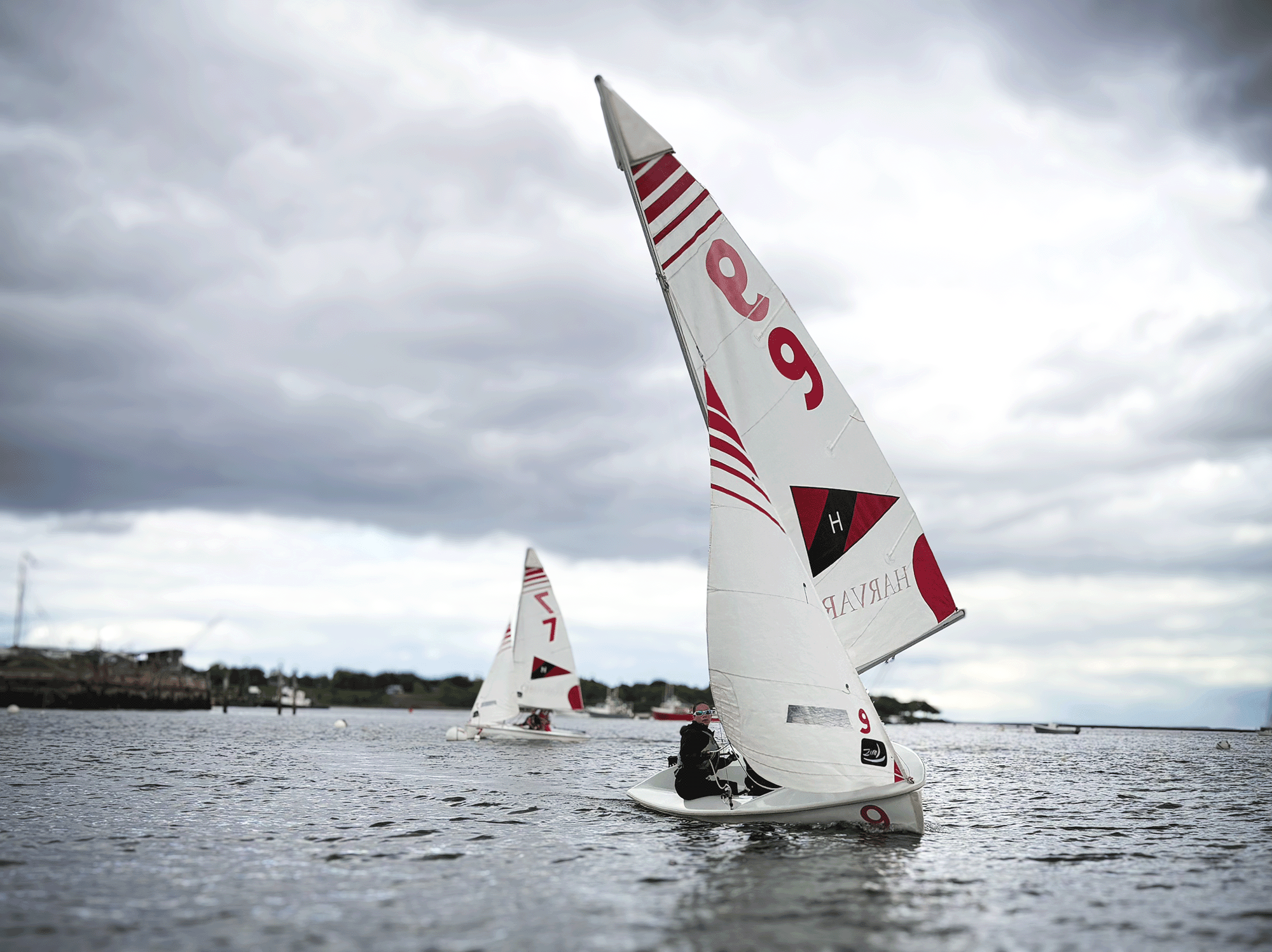 Sailing boats with sails on a body of water under a cloudy sky, with some people sailing and others nearby.
