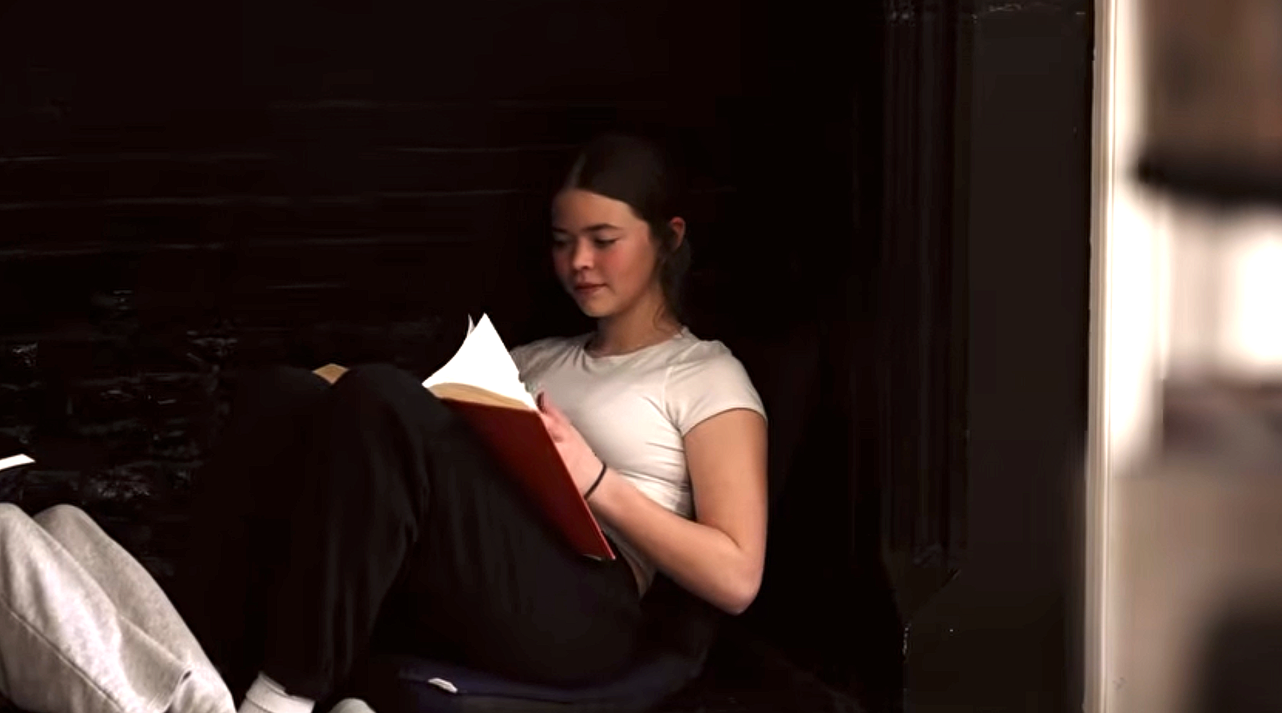 Young girl sitting on the floor, reading a book, with her legs crossed, inside a room with dark wood panel walls.