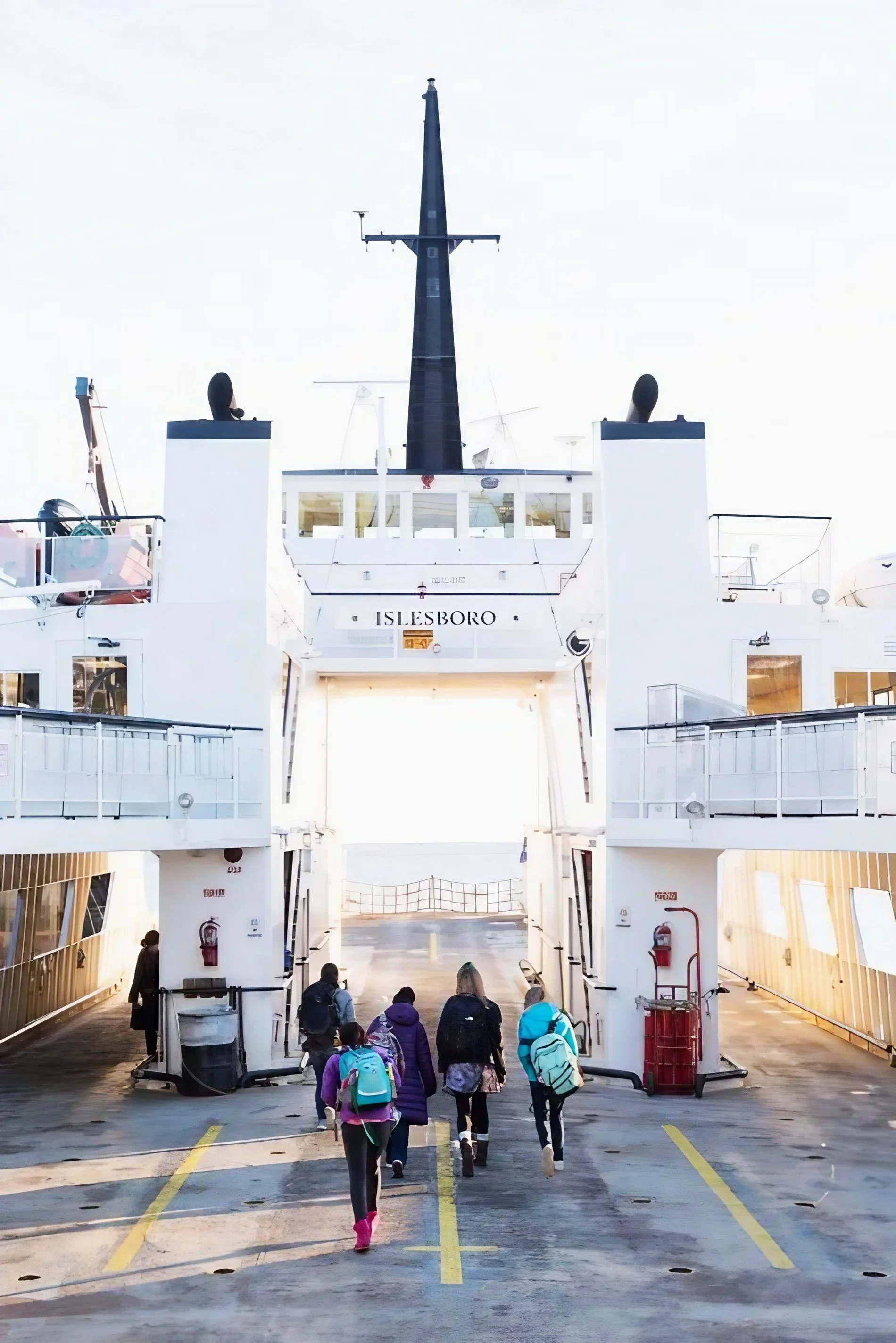 People walking on the deck of a cruise ship, approaching the entrance to the ship named ISLESBORO, with a large black ship horn and various antennas on top, and safety equipment on the sides.