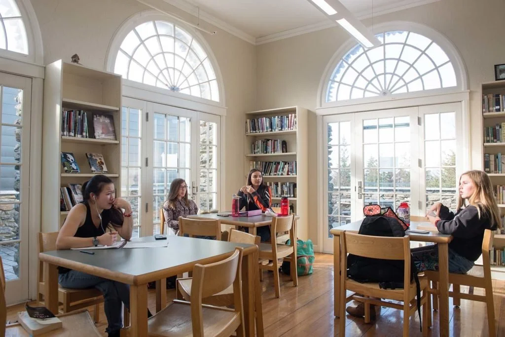Islesboro Central School students sitting at tables in a bright library room with large arched windows and bookshelves.