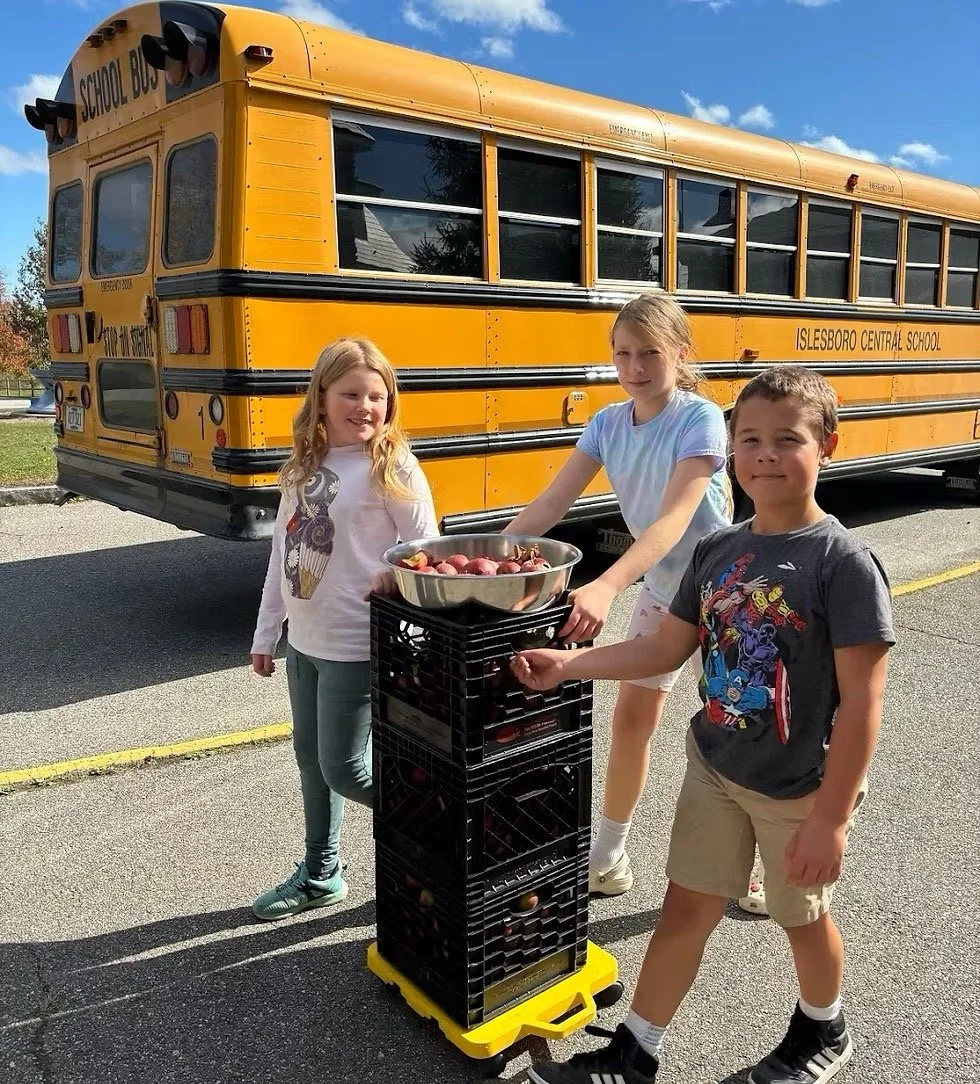 Three children near a yellow school bus, with two of them handling a crate of apples, in a parking lot on a sunny day.