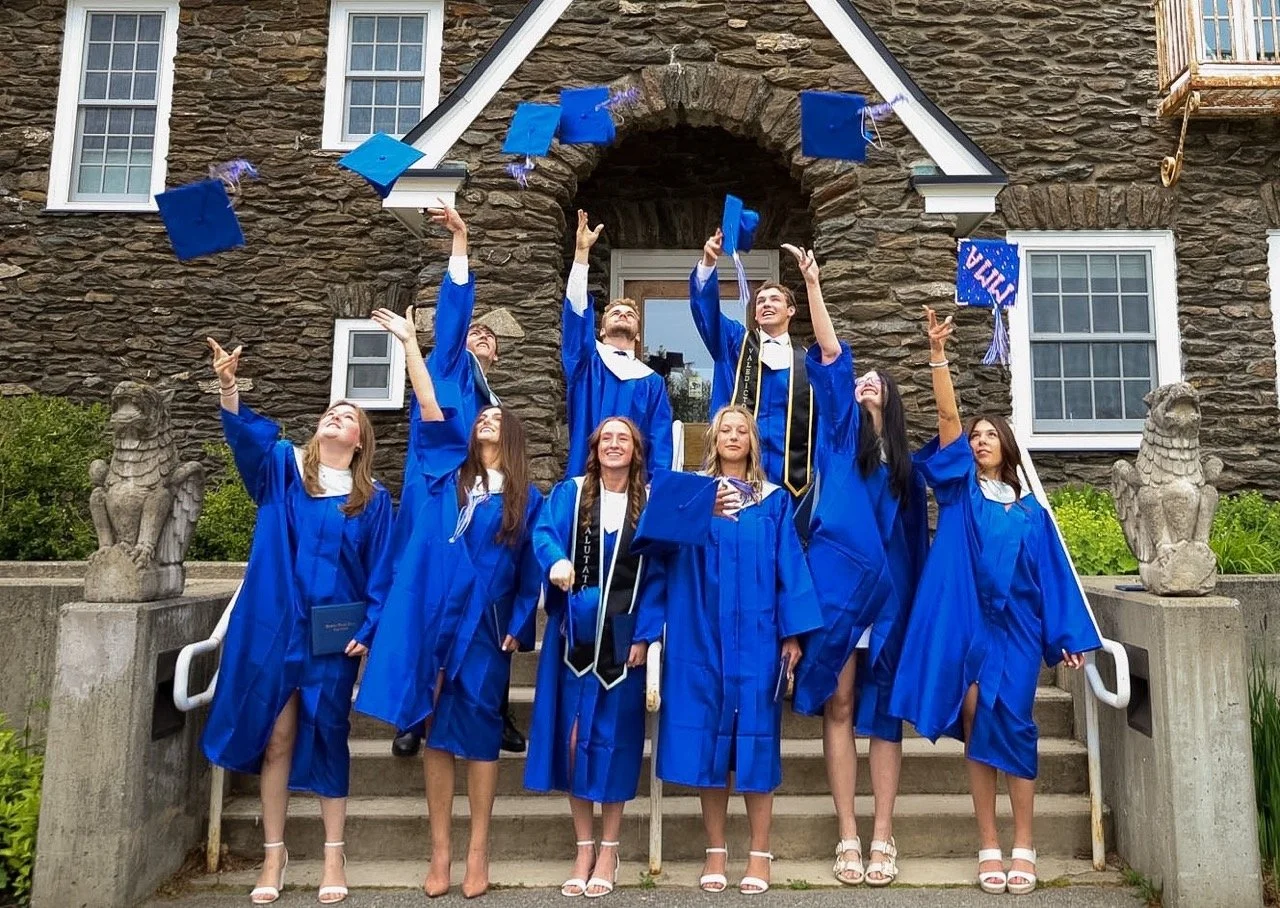 Graduates in blue caps and gowns celebrating by throwing their caps into the air on the steps of a stone building.
