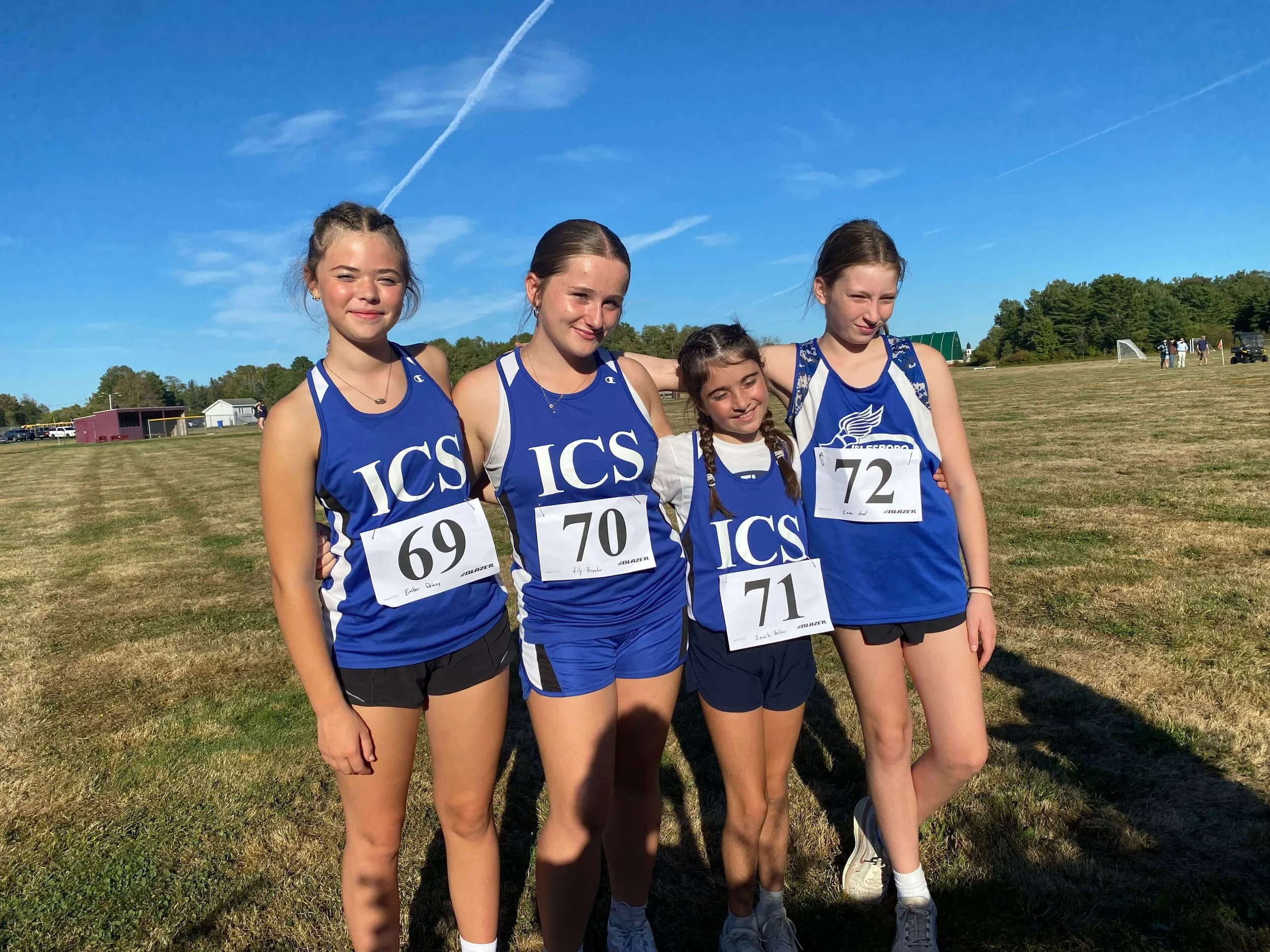 Four young girls in blue track uniforms standing outdoors on a sunny day, smiling, with a school field and other people in the background.