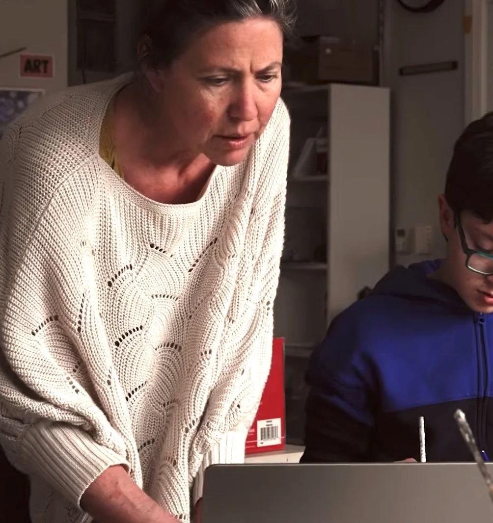 A woman with short brown hair leaning over a laptop, wearing a cream-colored knit sweater, in a room with bookshelves and art supplies in the background.