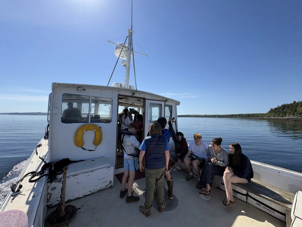Group of children and teenagers on a boat, some sitting on a bench and others standing near the cabin, with calm water and clear sky in the background.