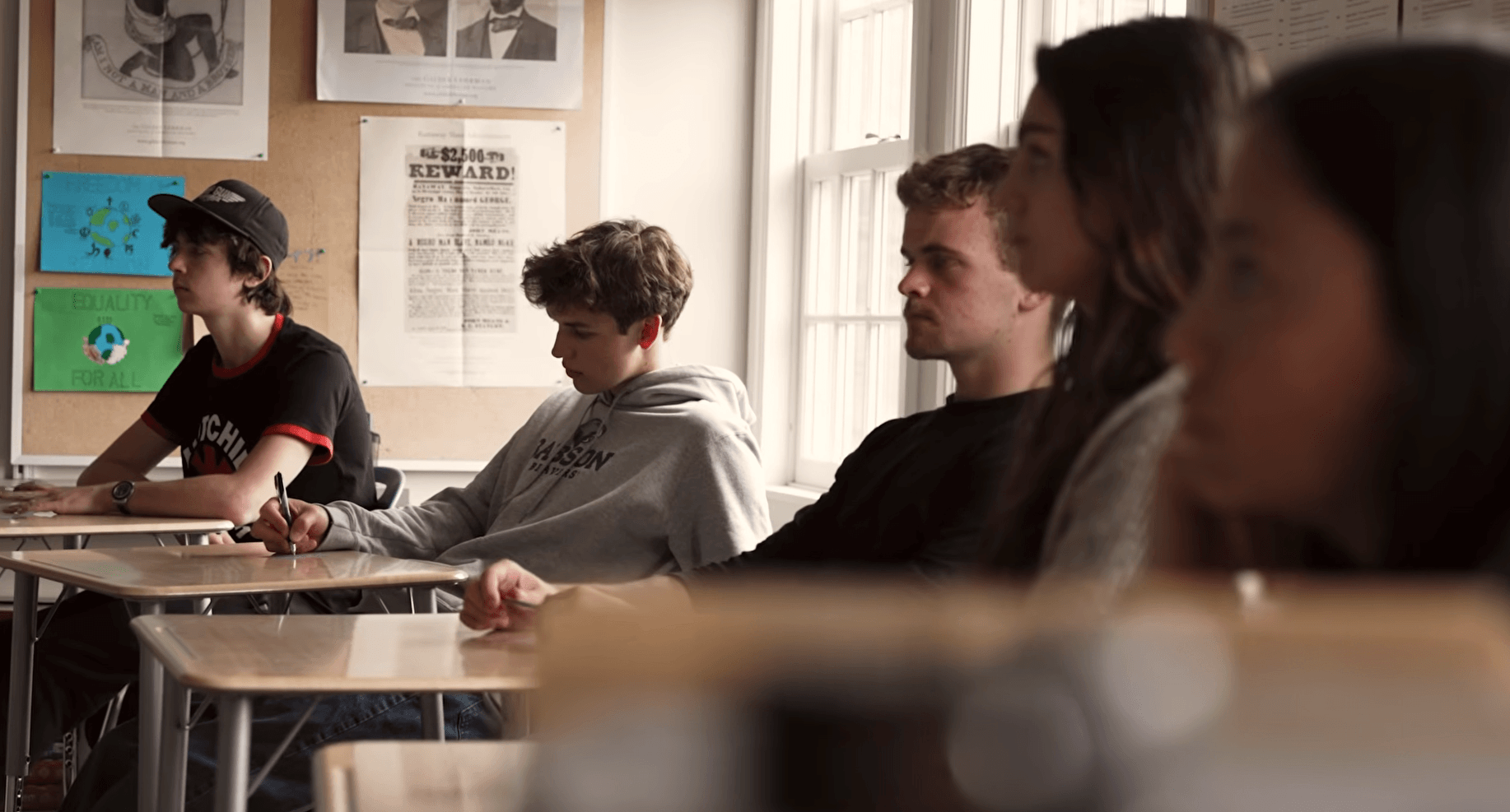 Students sitting at desks in a classroom, attentively listening during class, with bulletin boards and posters on the wall behind them.