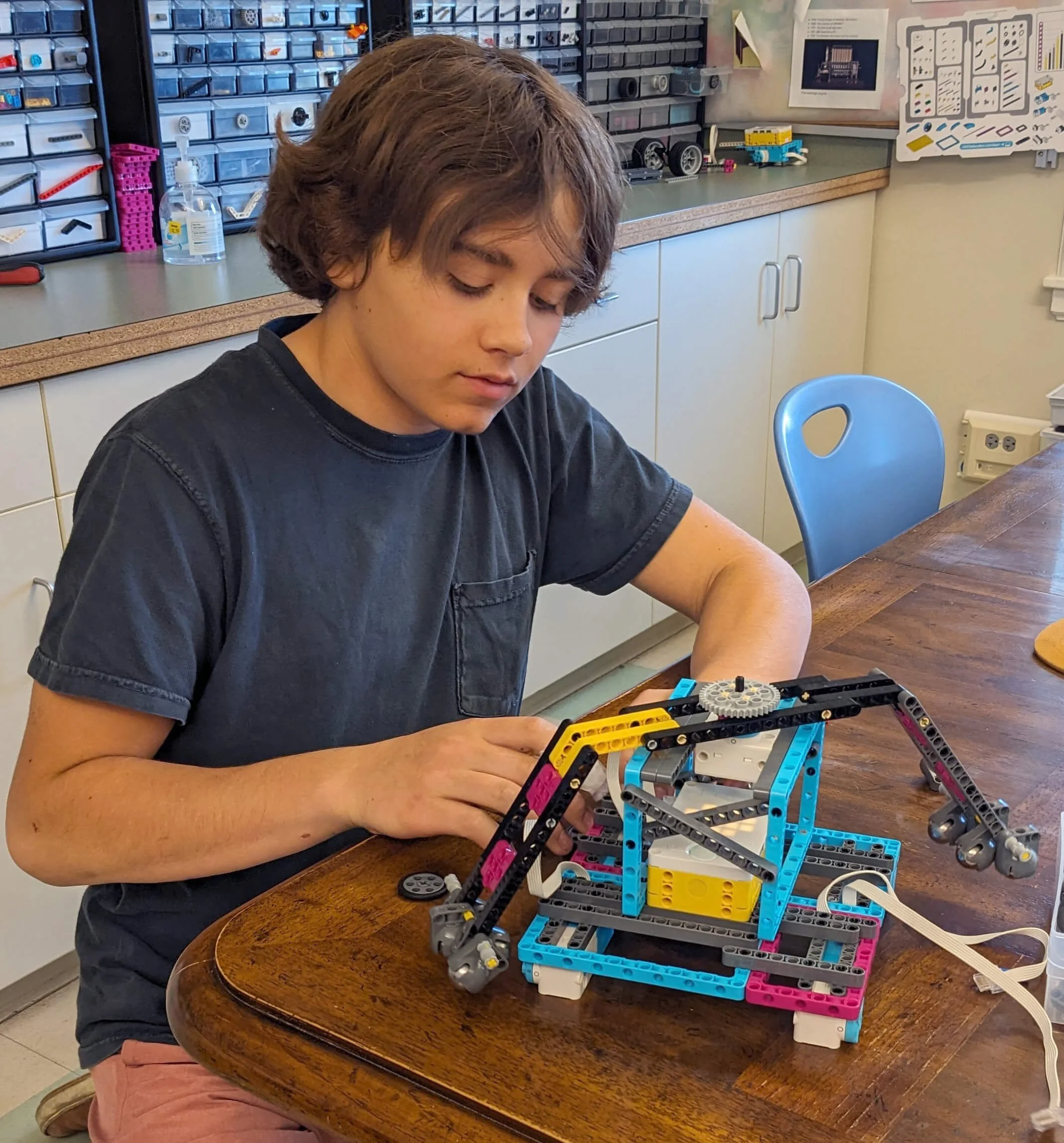 A young person sitting at a wooden table working on a robotic construction project made from colorful plastic parts and gears, with a background of storage bins filled with various robot components and tools.
