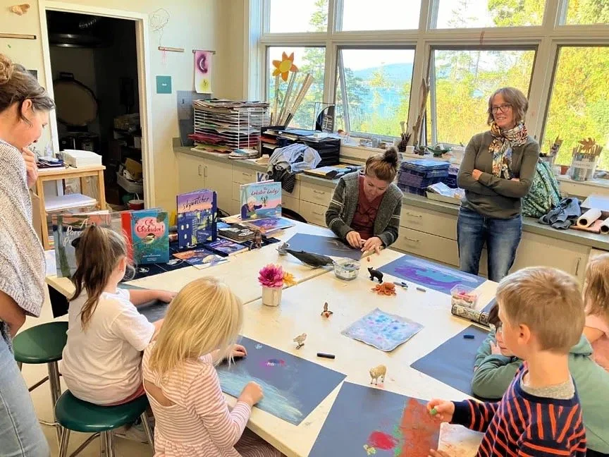 Children and adults gathered around a table in a classroom with books, art supplies, and decorations, engaged in art activities.