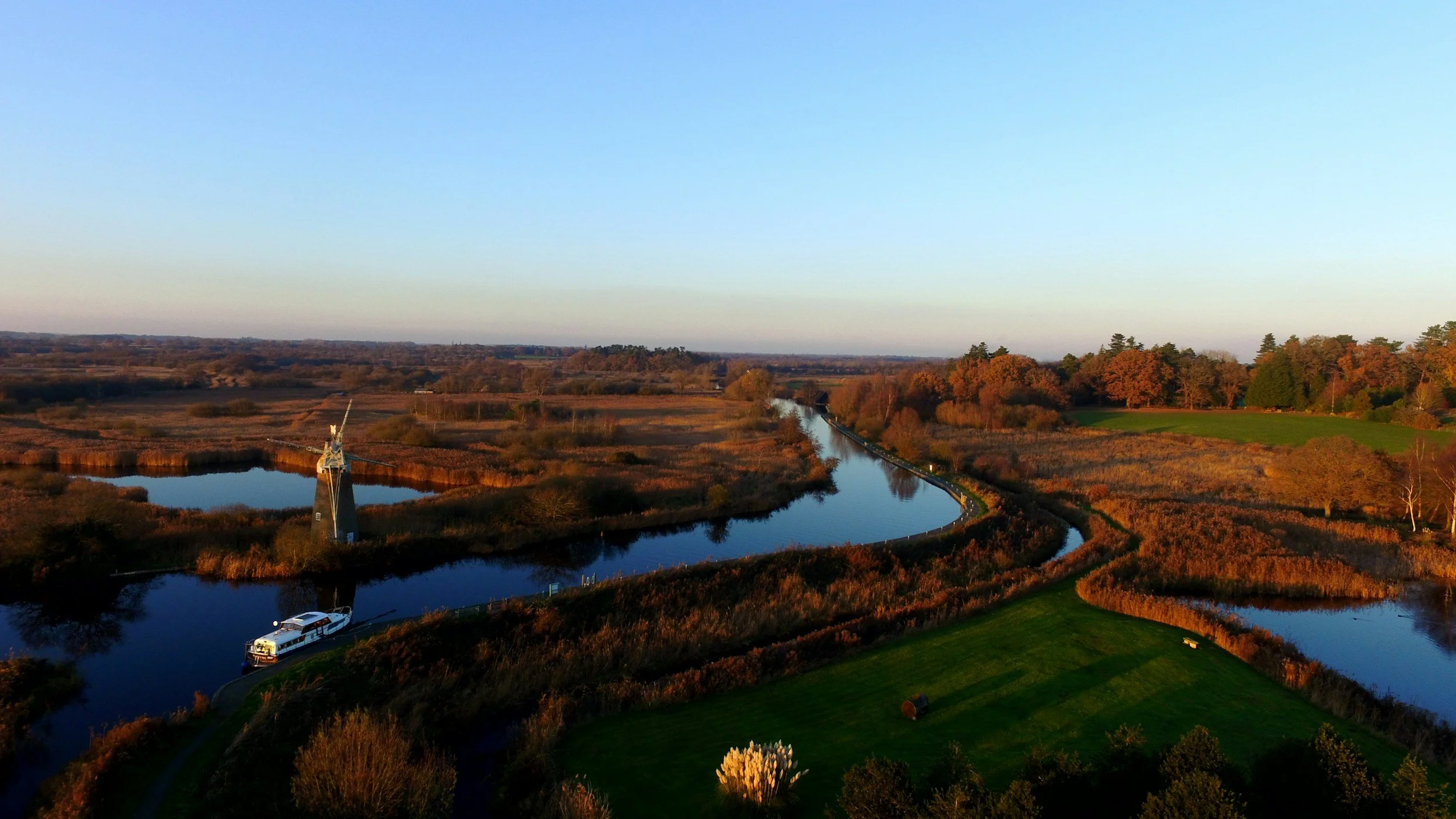 Aerial view of a rural landscape with winding water canals, a boat, windmill, trees, and open fields during autumn.