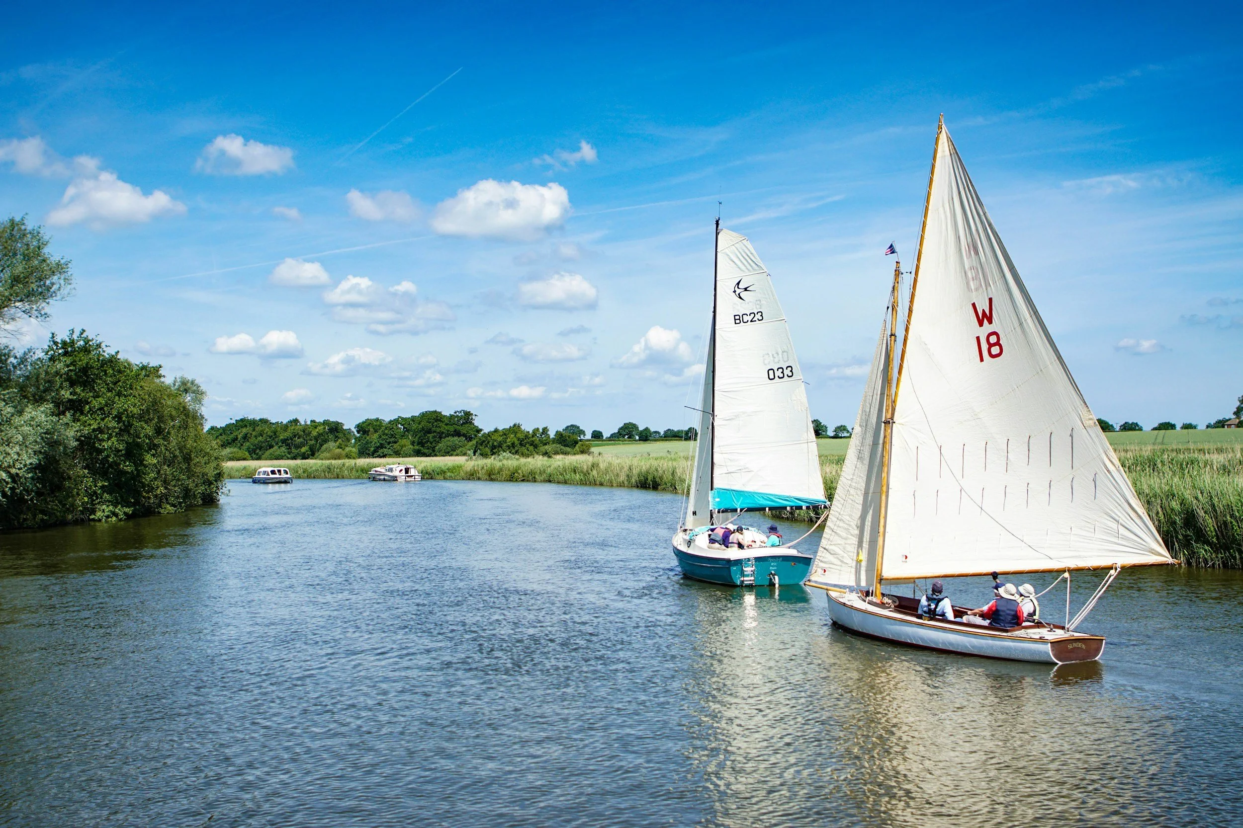 Sailboats with large white sails on a calm river, with a group of people on the boat, surrounded by green trees and a bright blue sky with scattered clouds.
