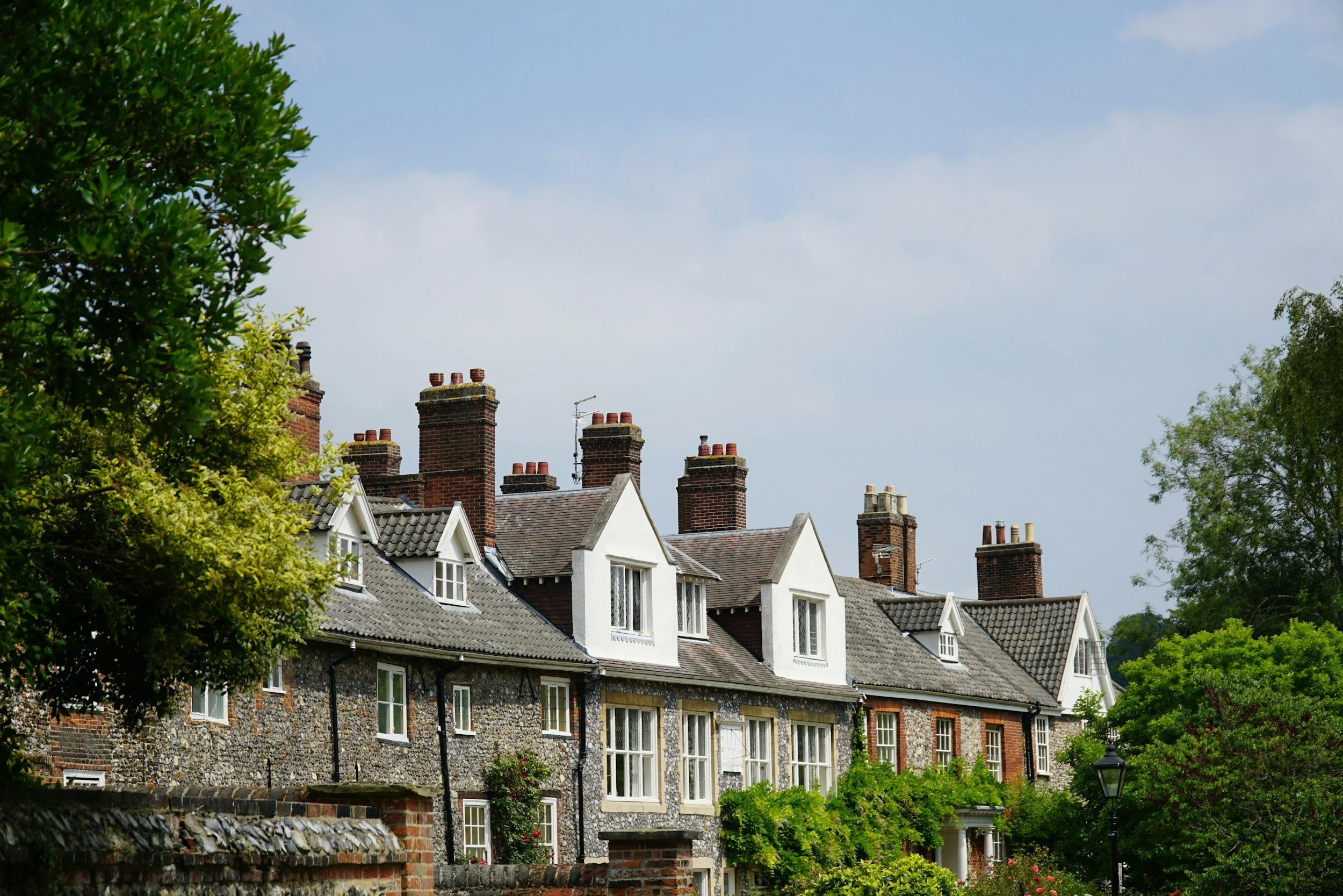 Row of houses with brick chimneys and slate roofs, partially obscured by green trees, under a blue sky with some clouds.