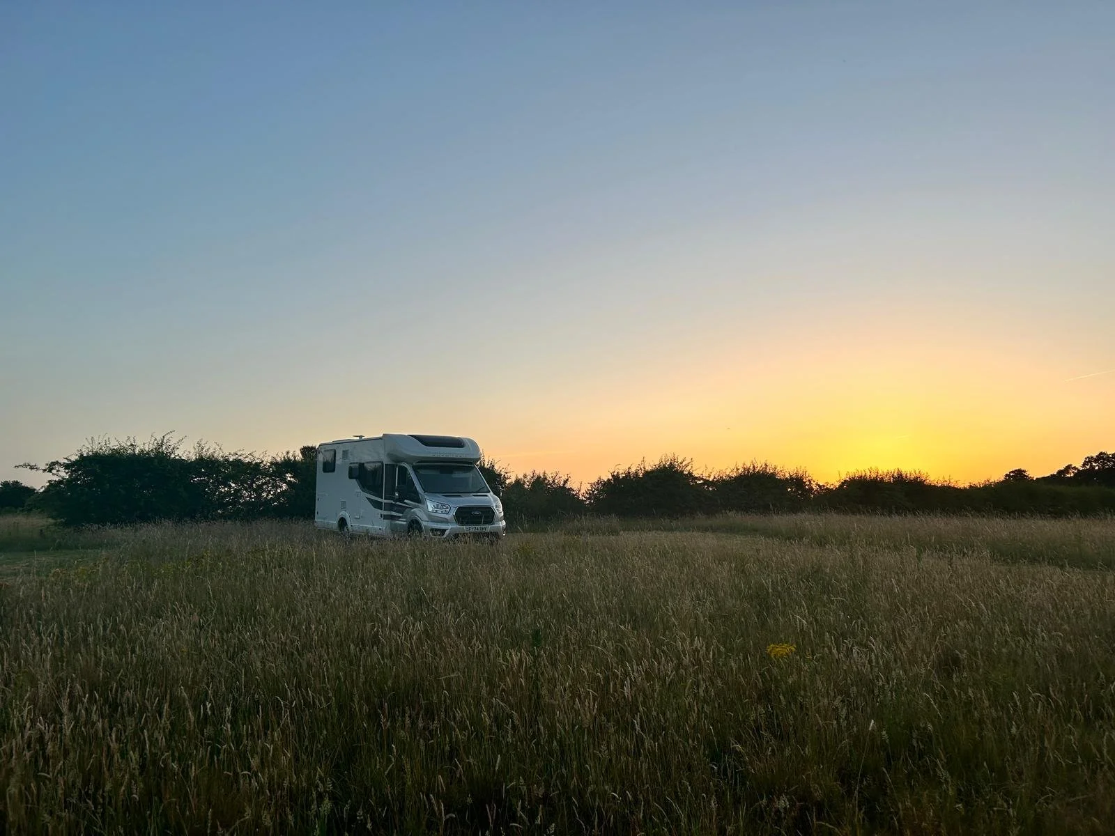 A white camper van parked in a grassy field during sunset with a clear sky and some trees in the background.