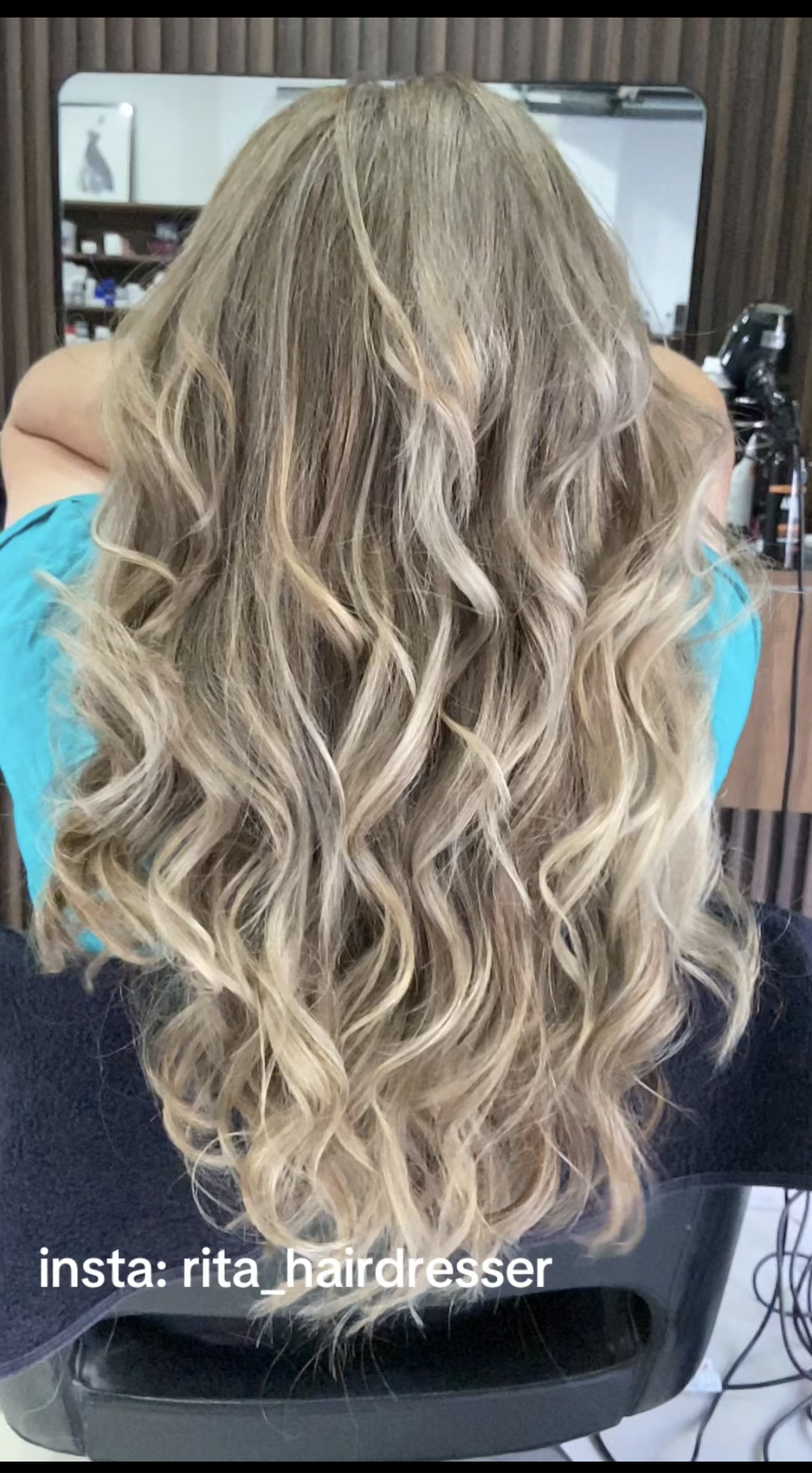 Woman with long, blonde, wavy hair sitting in a salon chair with a mirror and hairdressing tools in the background.