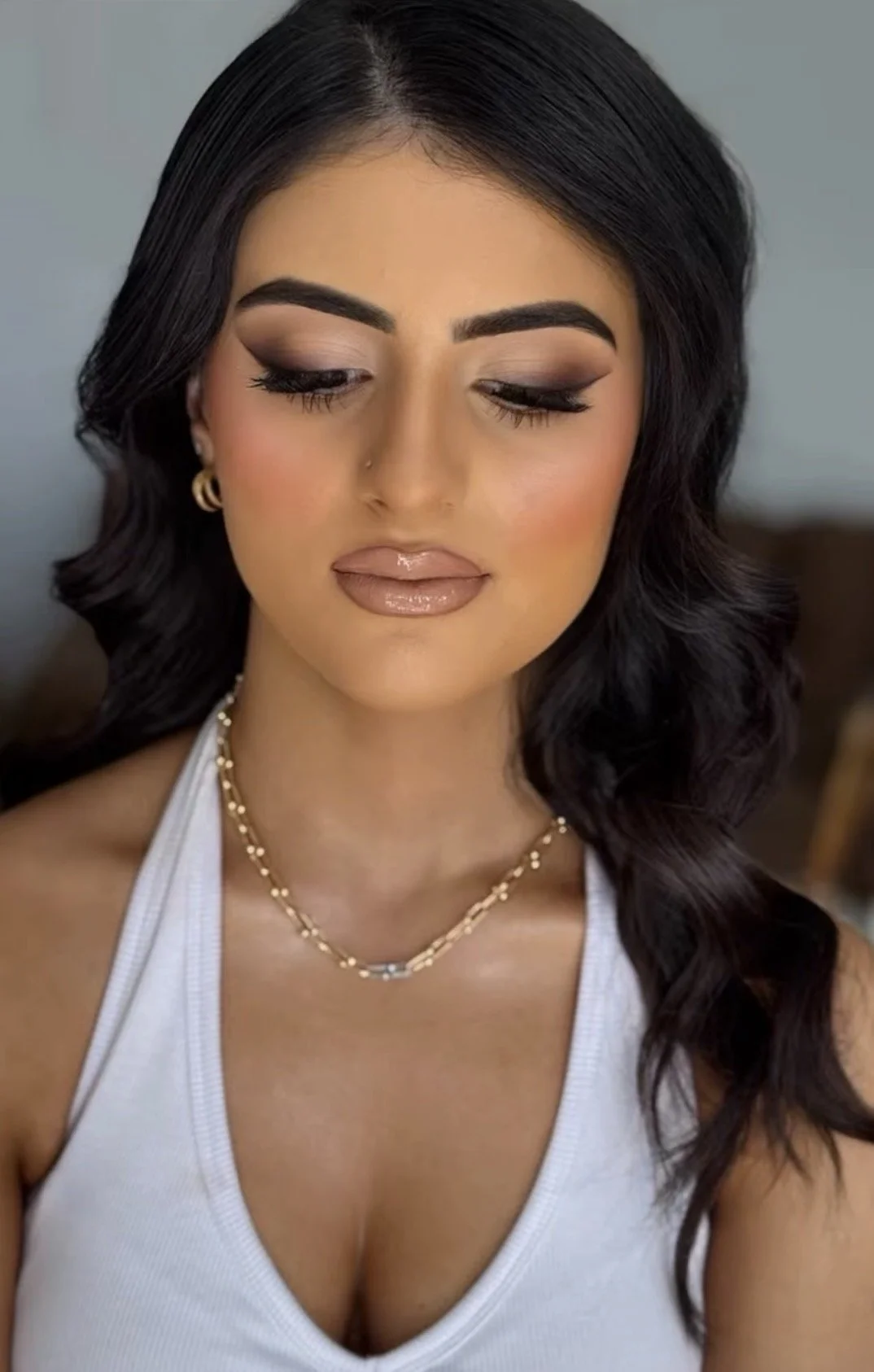 A woman with long black hair, wearing makeup, a white tank top, gold earrings, and a gold necklace, looking down with her eyes closed.