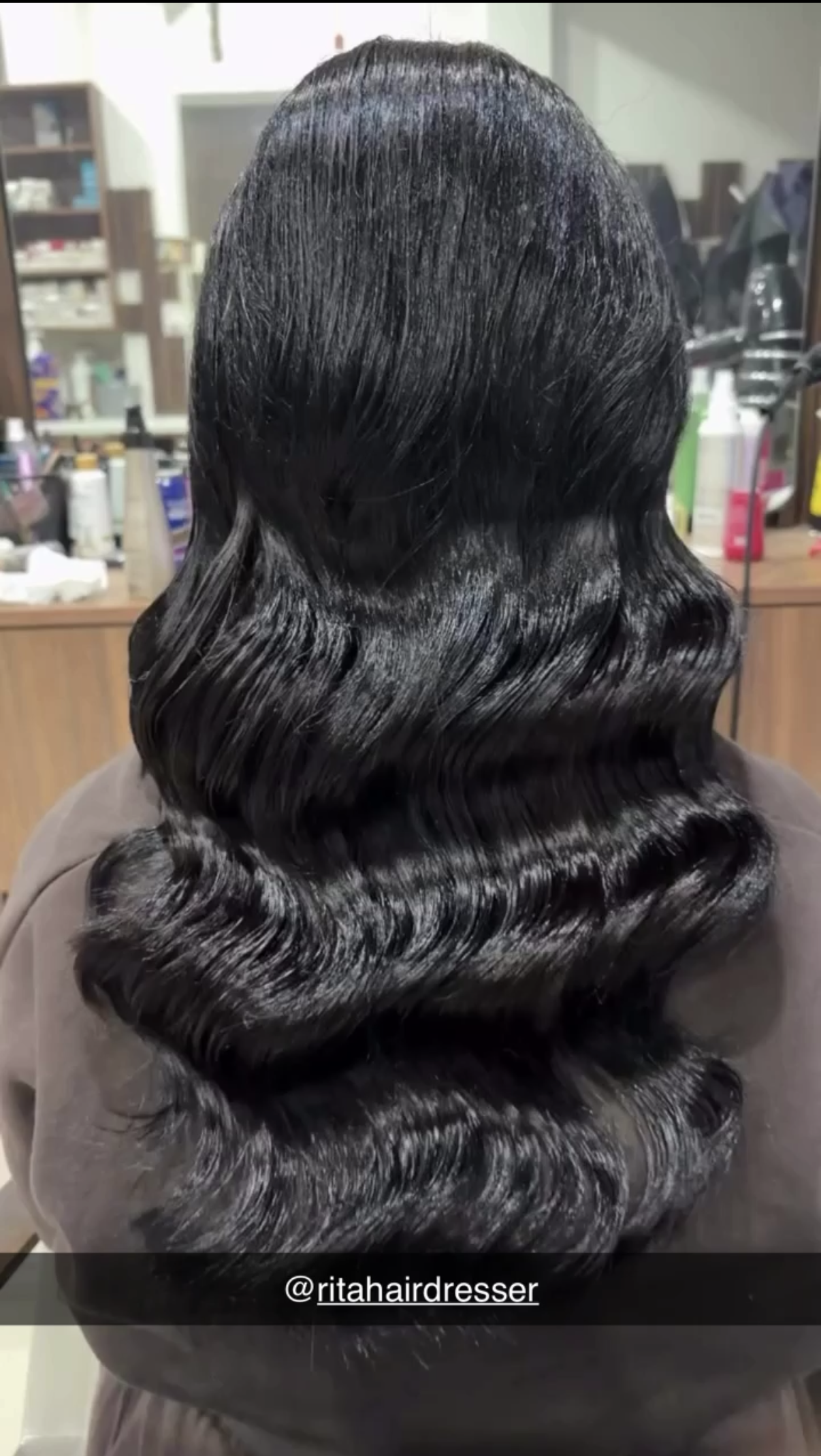Back view of a woman with long, glossy black hair styled in loose curls. In the background, beauty products and shelves are visible. The Instagram handle '@ritahairdresser' is at the bottom of the image.