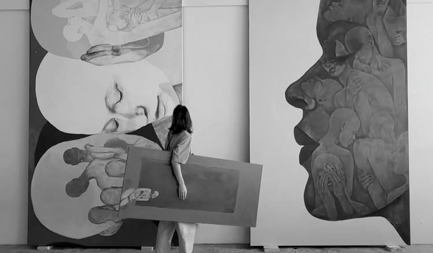 Een vrouw staat in een kunstgalerie met haar rug naar de camera, terwijl ze naar grote, monochrome schilderijen kijkt die menselijke gezichten en figuren afbeelden.