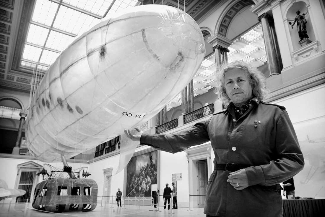 Man met lang, golvend haar en een jas met een striklint, poseert in een museum naast een groot luchtballon of blimp die hangt aan het plafond.