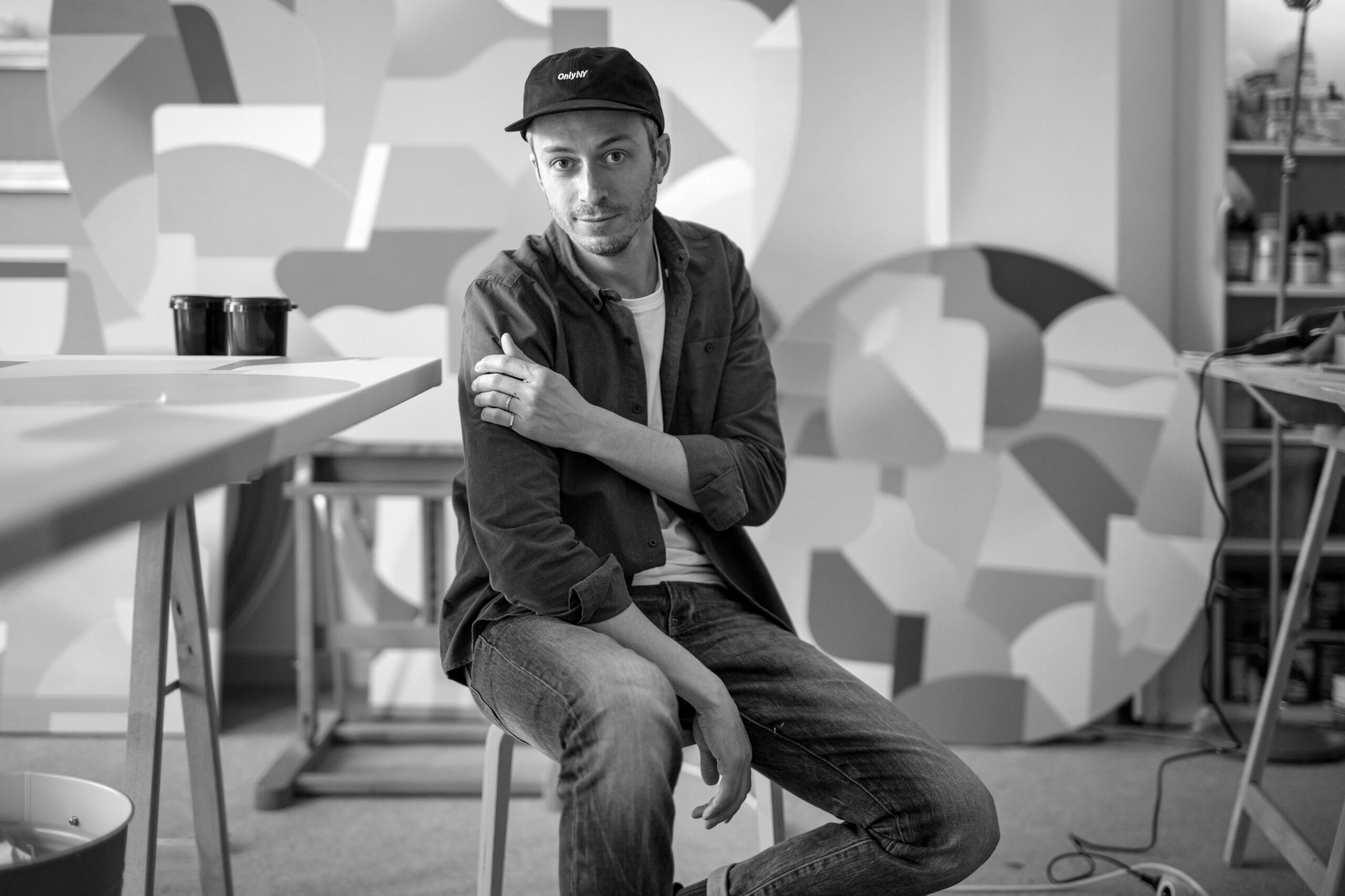 Een man zit op een stoel in een kunstatelier of kantoor, gekleed in een donker shirt en een pet, met een graffiti-achtige muur op de achtergrond.