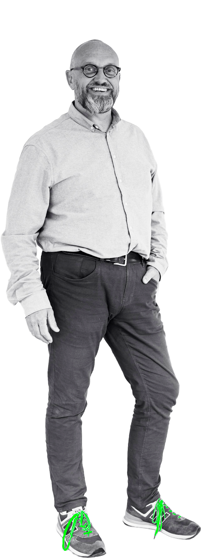 Man met een baard en bril, dragend een lichtblauw overhemd en donkere jeans, lachend en staand met een hand in de zak, in zwart-wit.