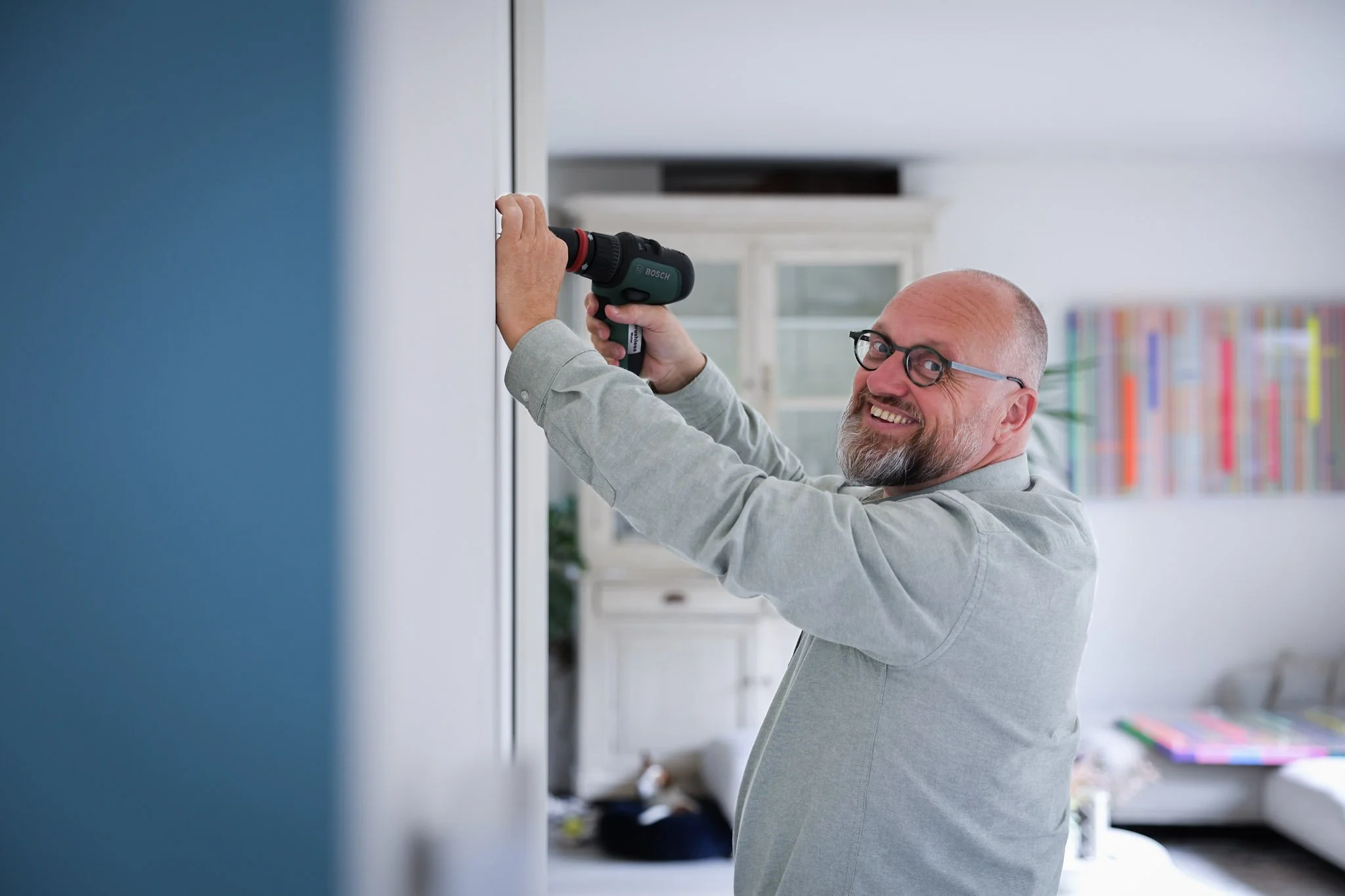 Man met een grijs overhemd gebruikt een boormachine om een muur in huis te boren.