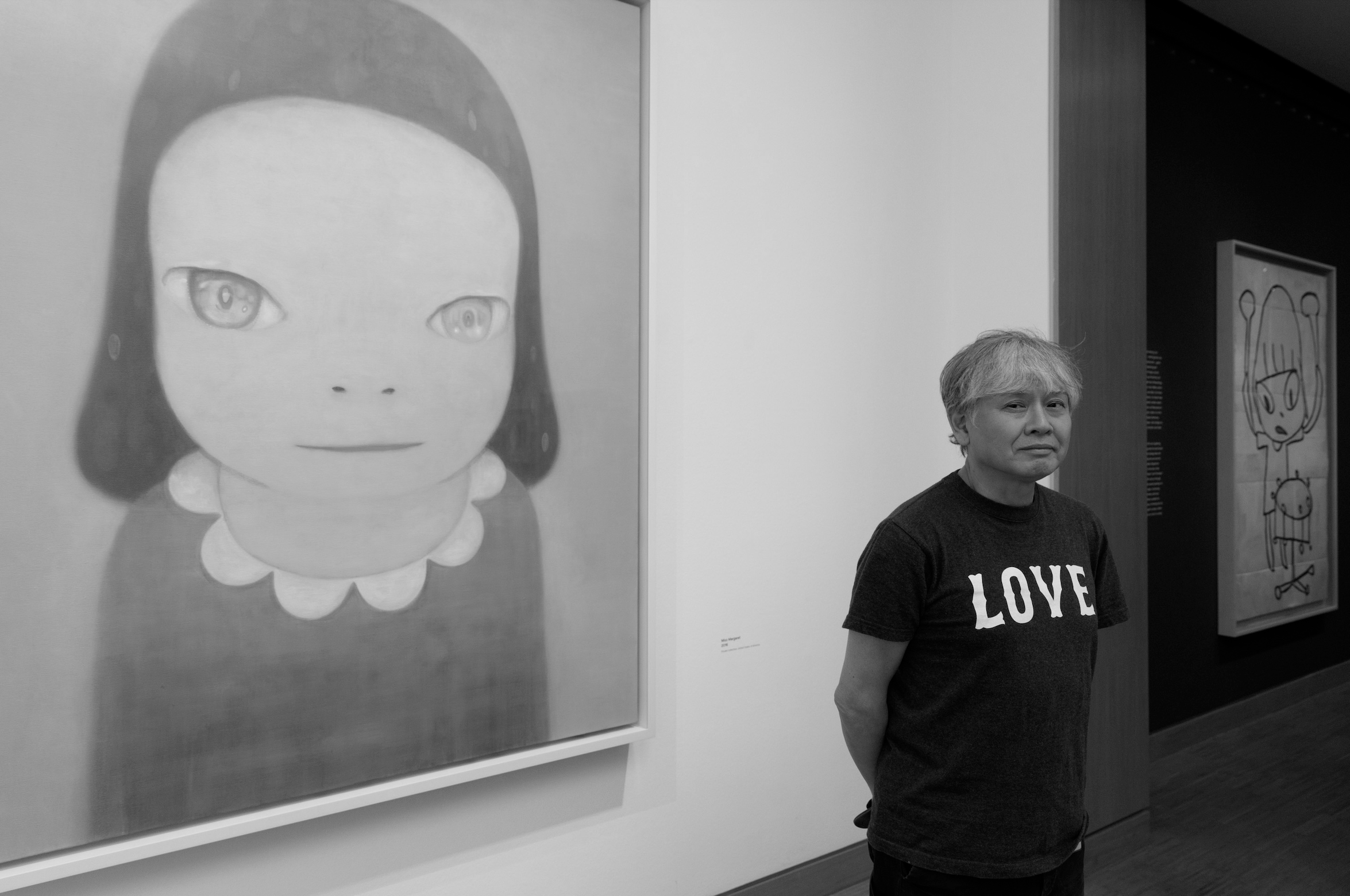 Man staat in een kunstmuseum voor twee kunstwerken, een grote tekening van een meisje met grote ogen en een andere eenvoudige tekening op de achtergrond. Hij draagt een zwart T-shirt met de tekst "LOVE".