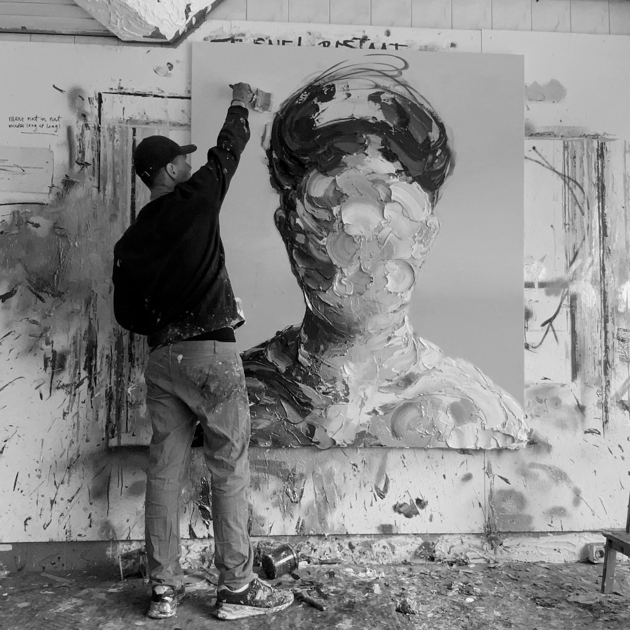 Een persoon schildert een groot zwart-wit abstract portret op een witte muur, omgeven door verfspatten en artistieke materialen.