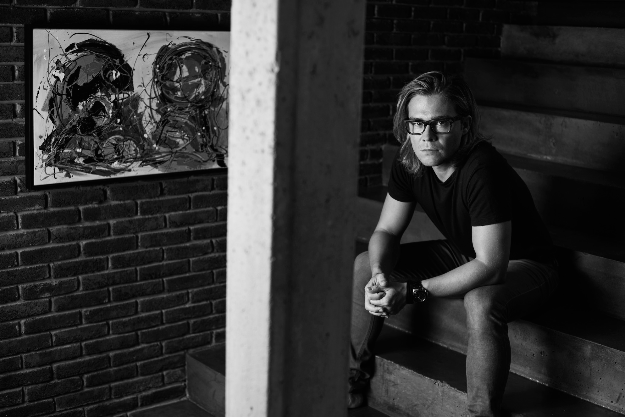een jonge man met bril zit op een trap in een industrieel gebouw, links een abstract zwart-wit schilderij aan de muur