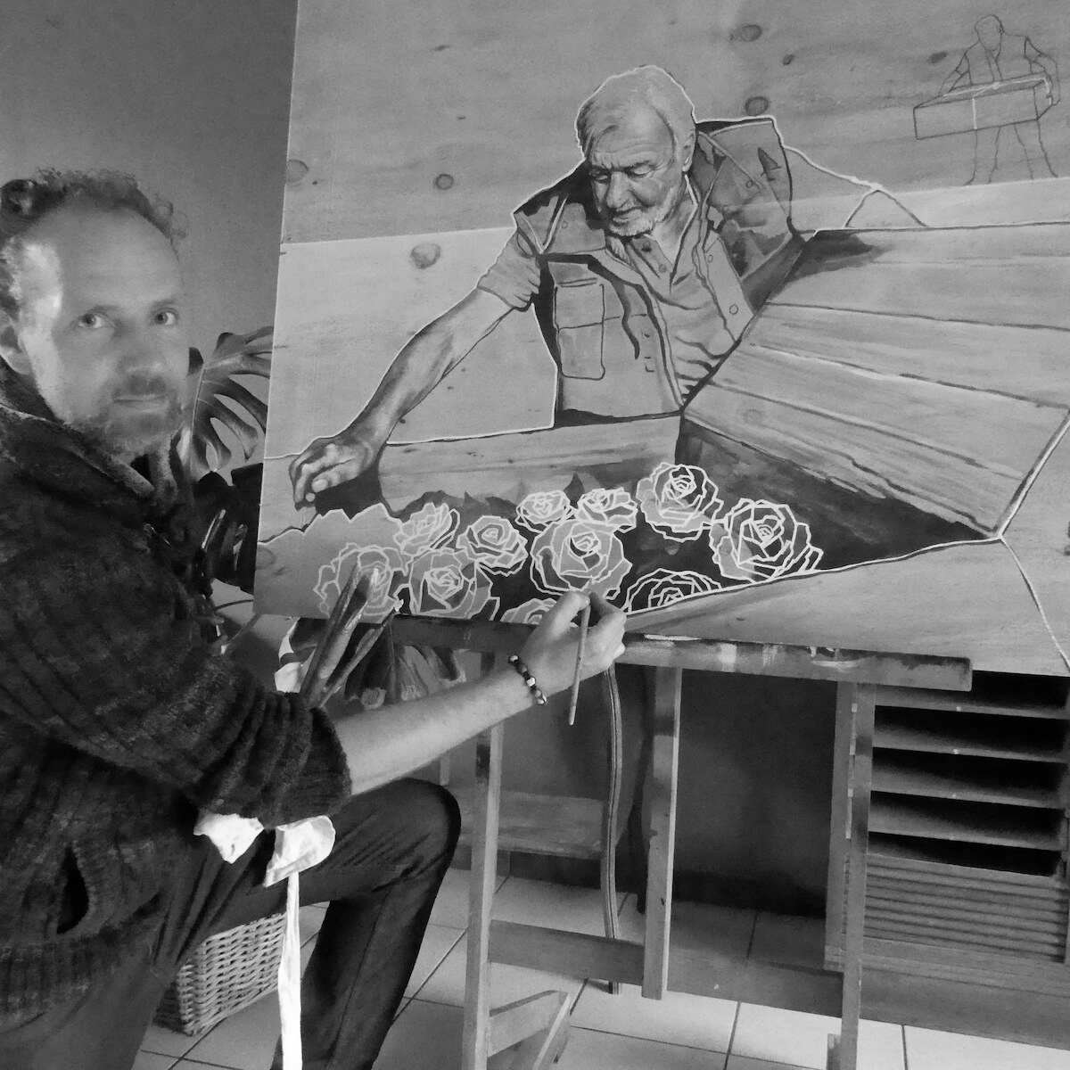 Een man schildert een zwart-wit portret op een groot paneel. Het portret toont een oudere man met een baard die naar beneden kijkt, met bloemen voor hem. De schilder is bezig met het toevoegen van details.