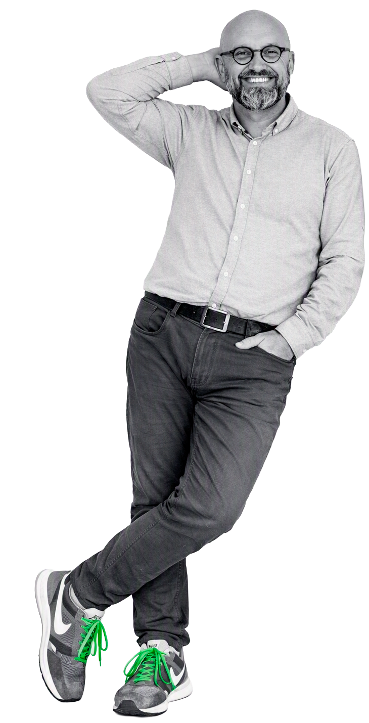 Man met bril, glimlach, korte baard, in grijs overhemd en zwarte jeans, staand met arm achter hoofd en andere hand in zak, op sneakers met groene veters.