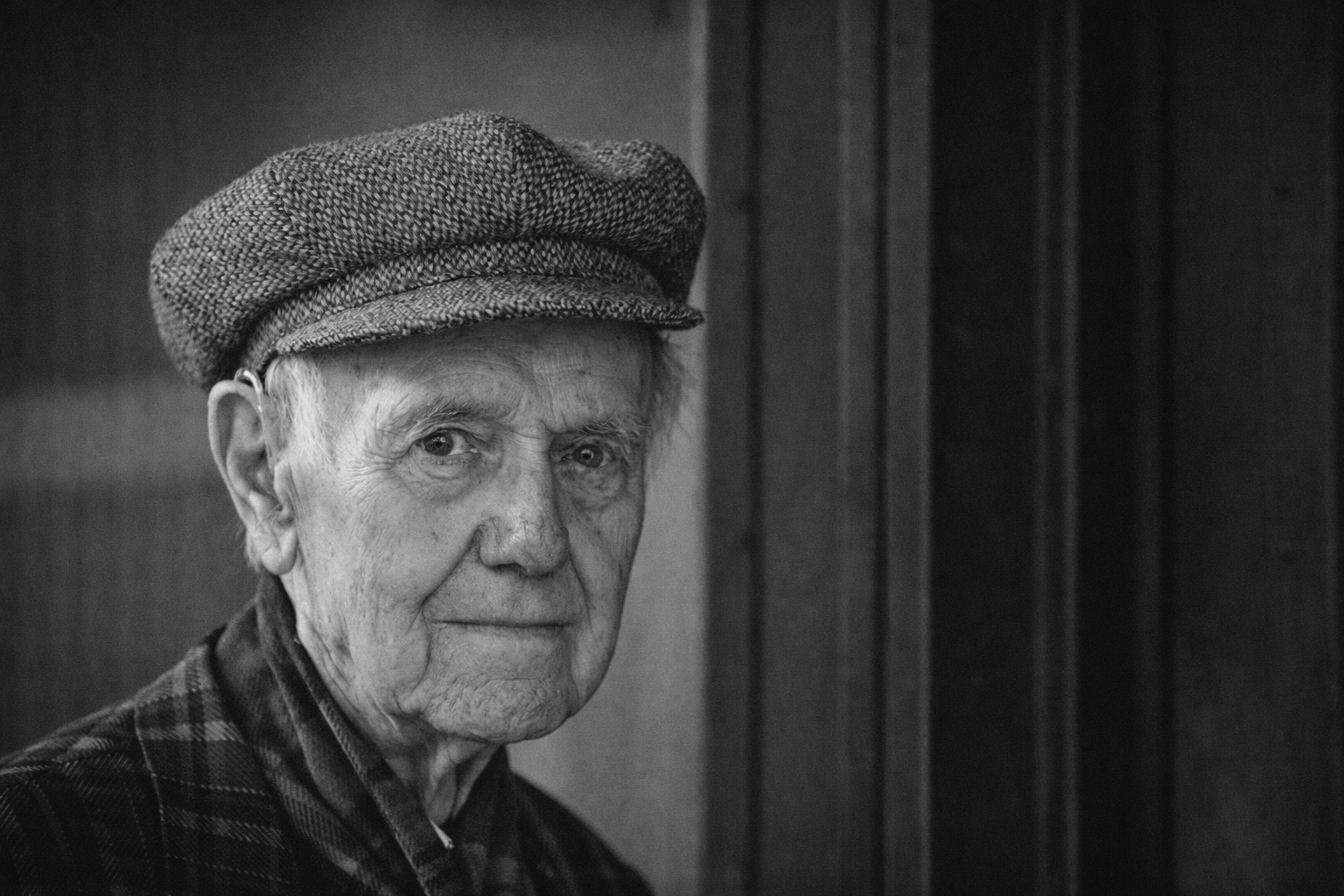 Zwart-wit foto van een oude man met een pet en een chequered jas, met een achtergrond van houten panelen.