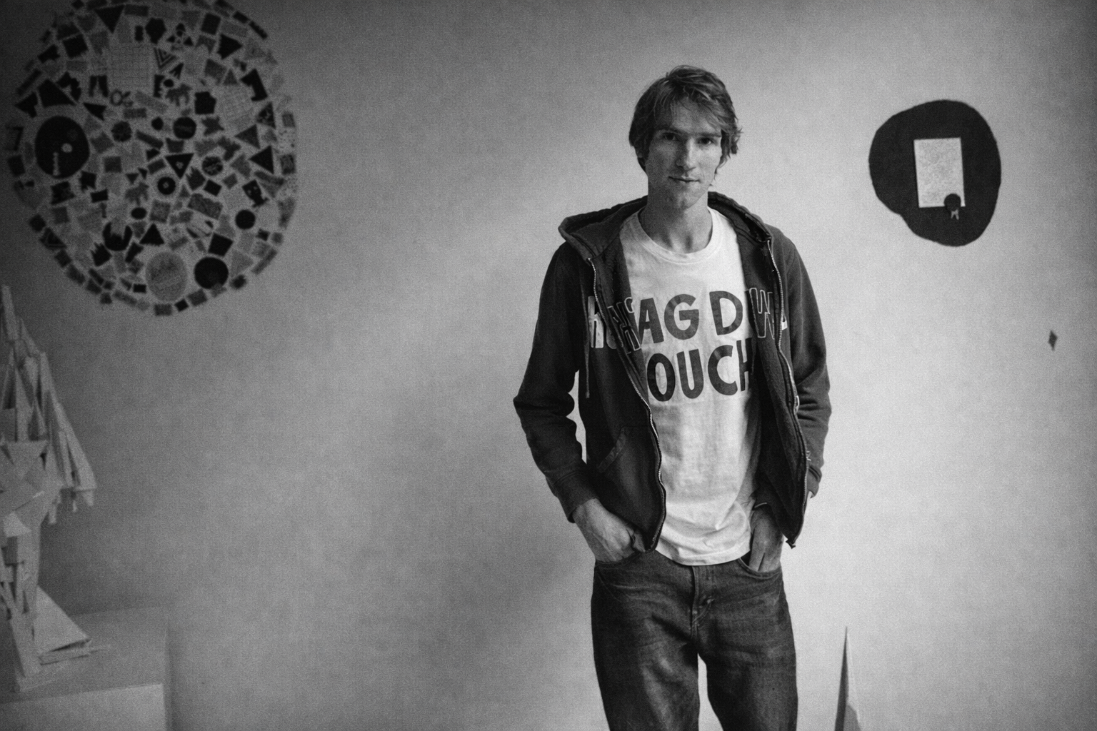 Een jonge man met een krullende, middelmatige lengte haar staat voor een muur met grafische kunst. Hij draagt een witte T-shirt met zwarte tekst en een zwarte jas, met beide handen in zijn zakken.