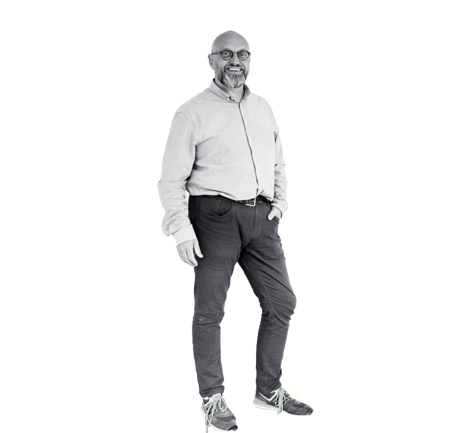 Een lachende man met een baard en bril, gekleed in een lichtgekleurde overhemd en donkergekleurde broek, staand met één hand in de zak en een grote witte achtergrond.