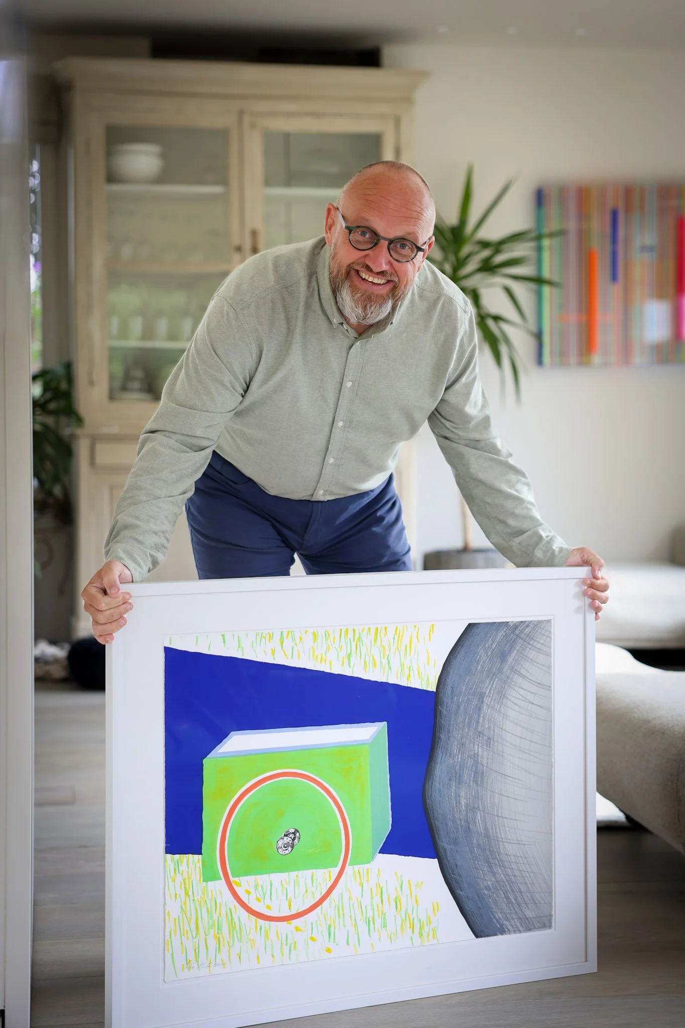 Een man met een grijze baard en ronde bril die glimlacht, terwijl hij een kunstwerk vasthoudt in een woonkamer. Op de achtergrond een houten kast met servies en een kleurrijk schilderij aan de muur.