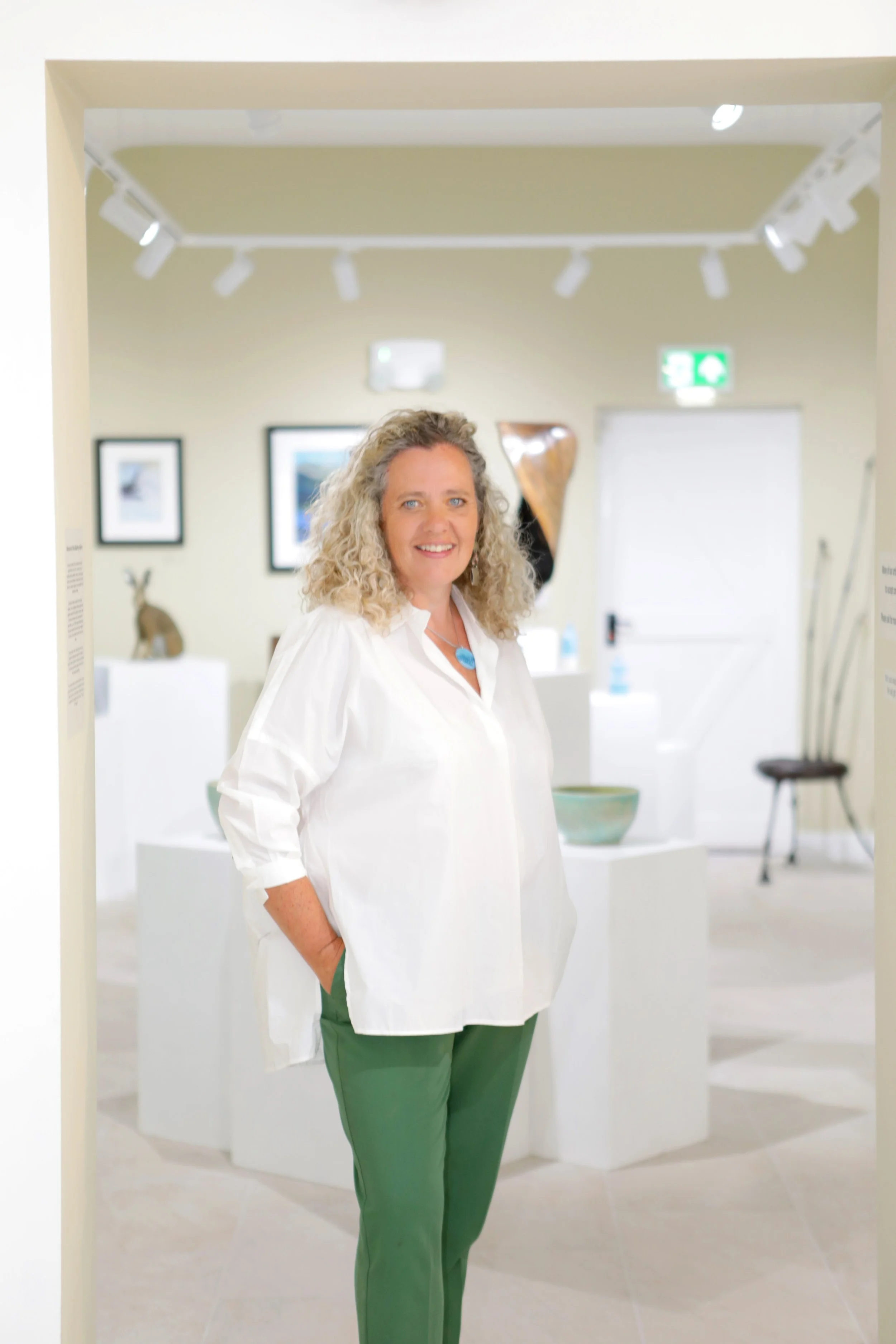 A woman with curly blonde hair smiling, wearing a white blouse and green pants, standing in an art gallery with framed artwork and sculptures.