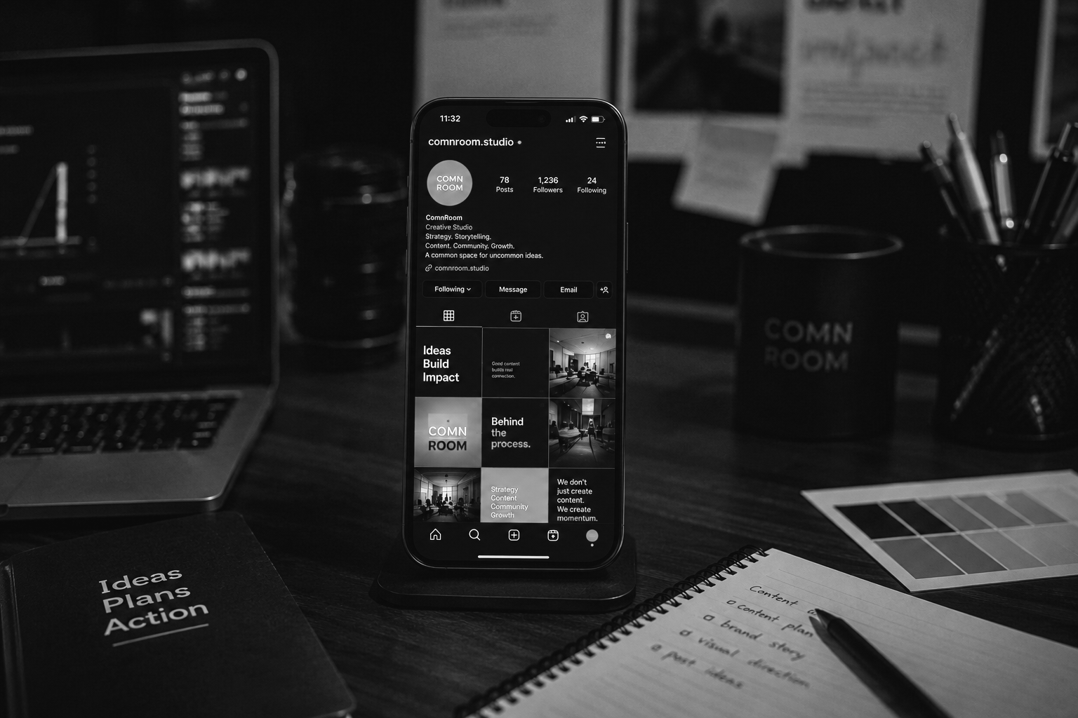 Black and white photo of a workspace with a laptop, a smartphone displaying an Instagram profile of 'comnroom.studio', a notebook, and various office supplies on a wooden desk.