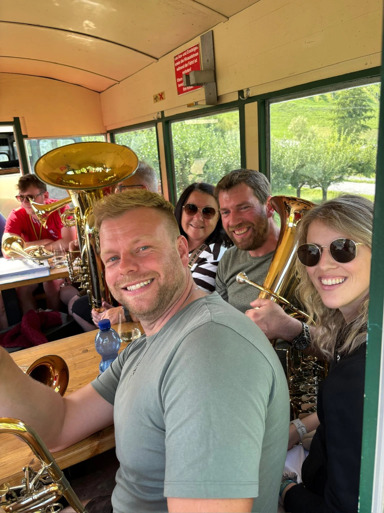 Junge Männer und Frauen mit Blasinstrumenten in einem Raum mit großen Fenstern und grüner Landschaft im Hintergrund.