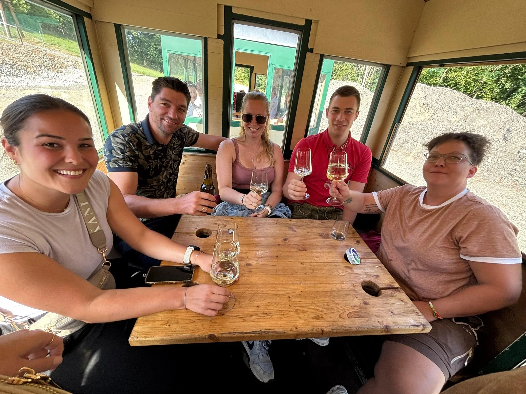 Fünf Menschen sitzen im Inneren eines Holzhauses, lachen und trinken Wein. Im Hintergrund sind große Fenster mit Blick auf eine grüne Landschaft.