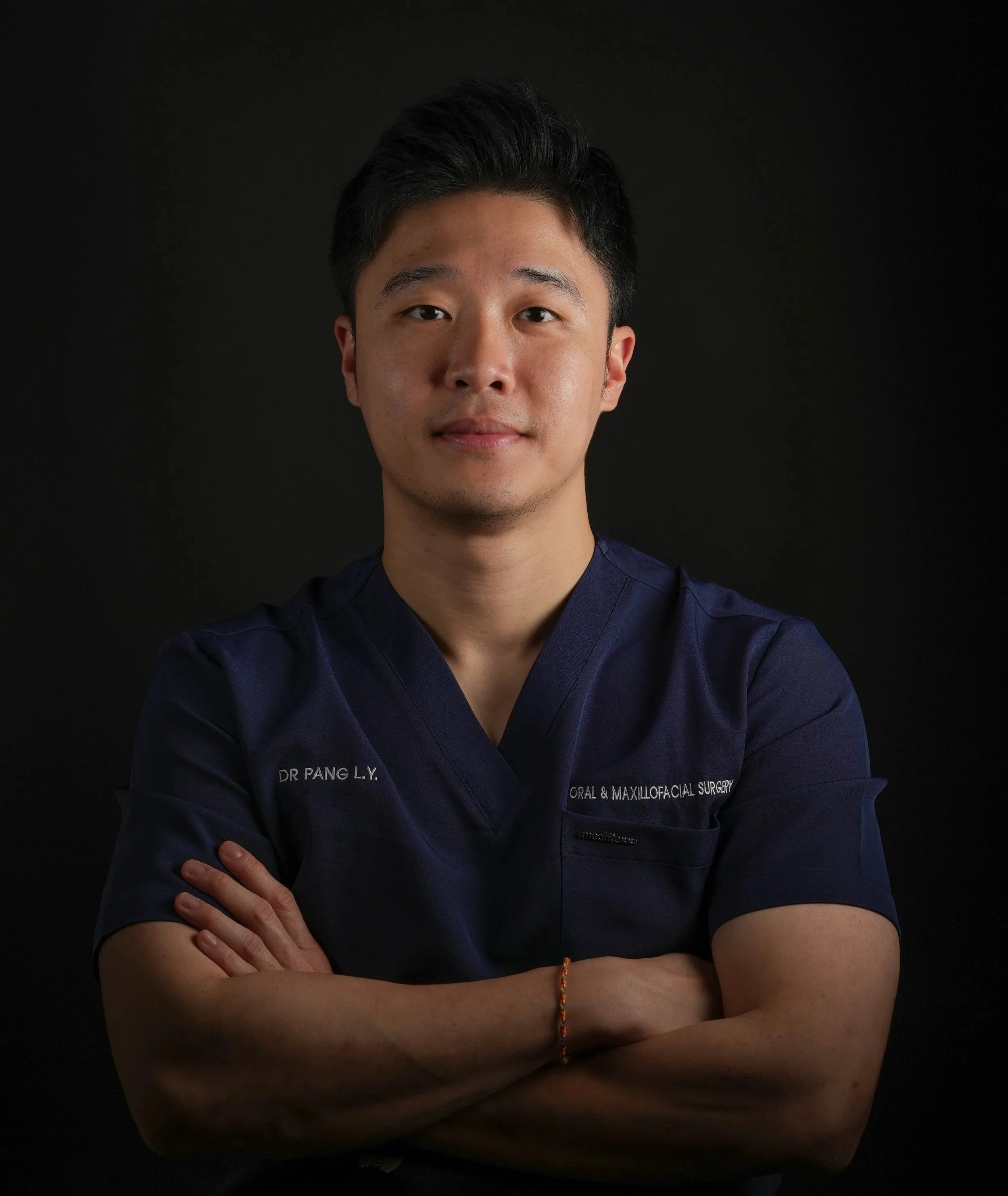 A young man with black hair wearing a navy medical uniform with the name tag 'Dr. Pang L.Y.' and the text 'Oral & Maxillofacial Surgery,' posing with crossed arms against a solid black background.