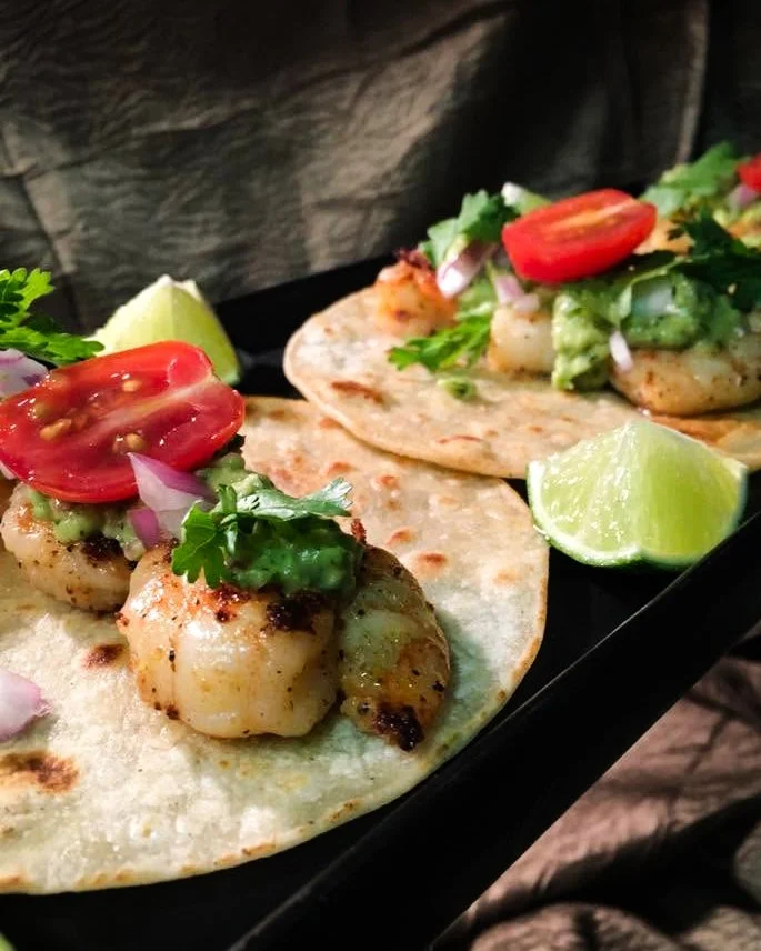 Two fish tacos topped with cherry tomatoes, chopped onions, cilantro, and guacamole, served with lime wedges.