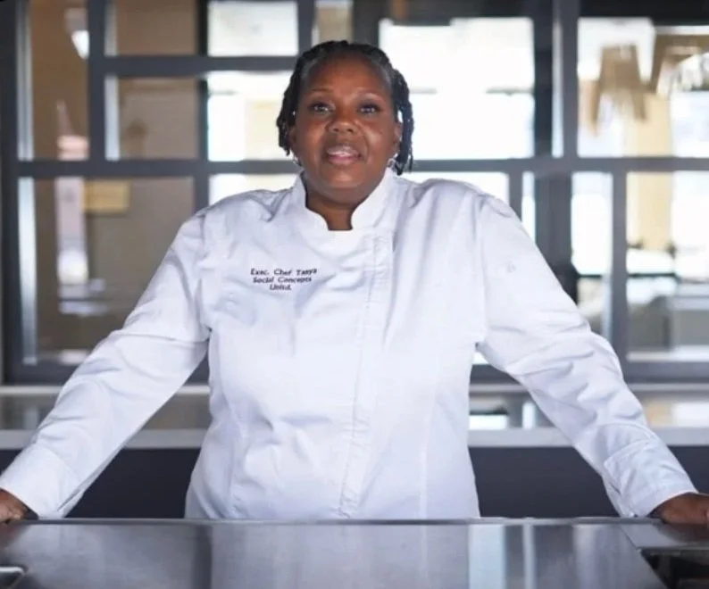 A woman dressed in a white chef's coat standing behind a kitchen counter in a commercial kitchen environment.