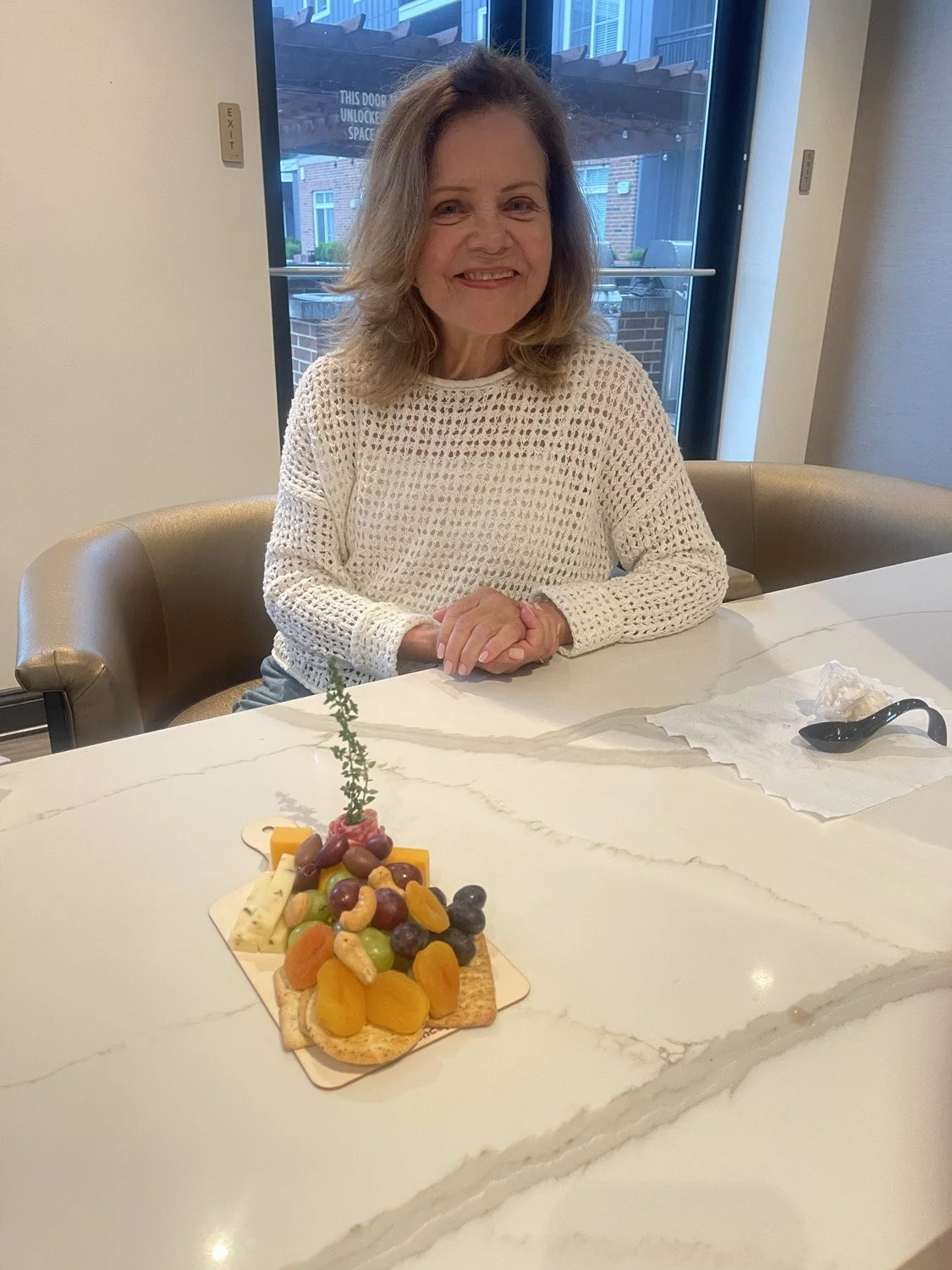 A woman sitting at a table with a cheese and fruit platter, smiling, with a window in the background showing a brick building and patio.