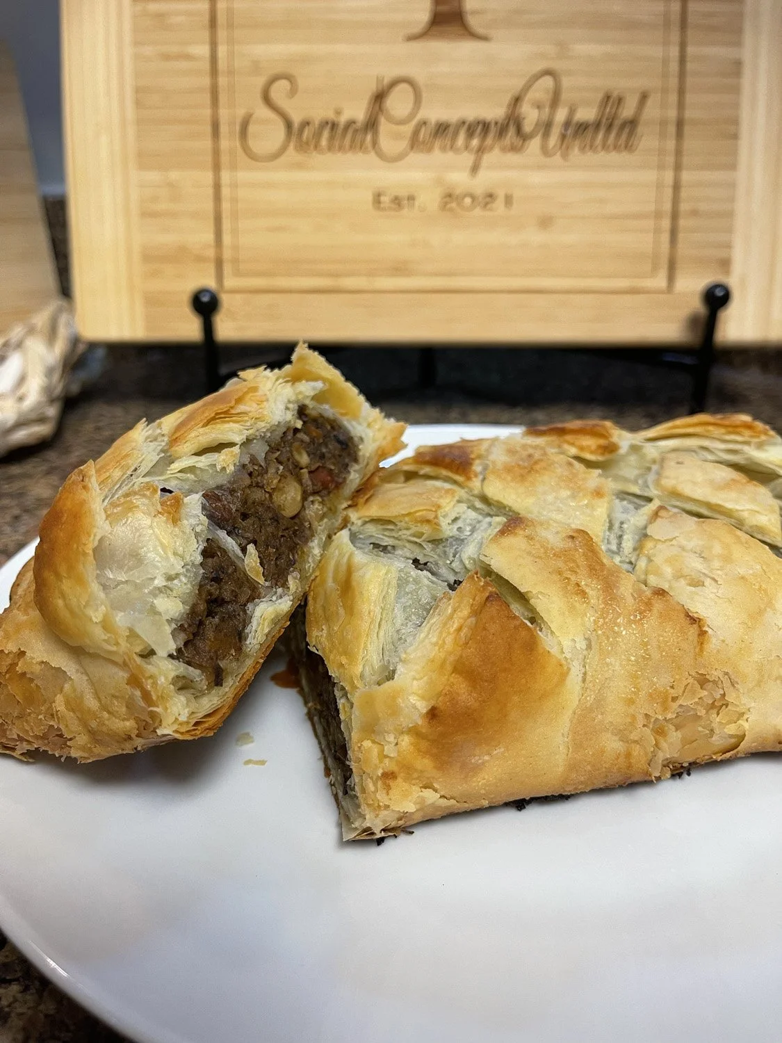 A plate with sliced savory meat pie with a flaky golden crust, showcasing a dark meat filling with vegetables inside, on a white plate. In the background, there is a wooden sign with the text 'Social Concept Wealth Est. 2021'.