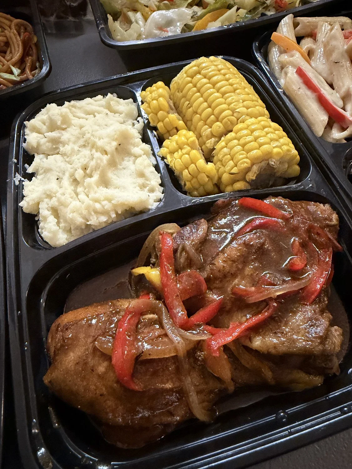 A meal tray with mashed potatoes, corn on the cob, and a meat dish with red bell peppers in gravy.