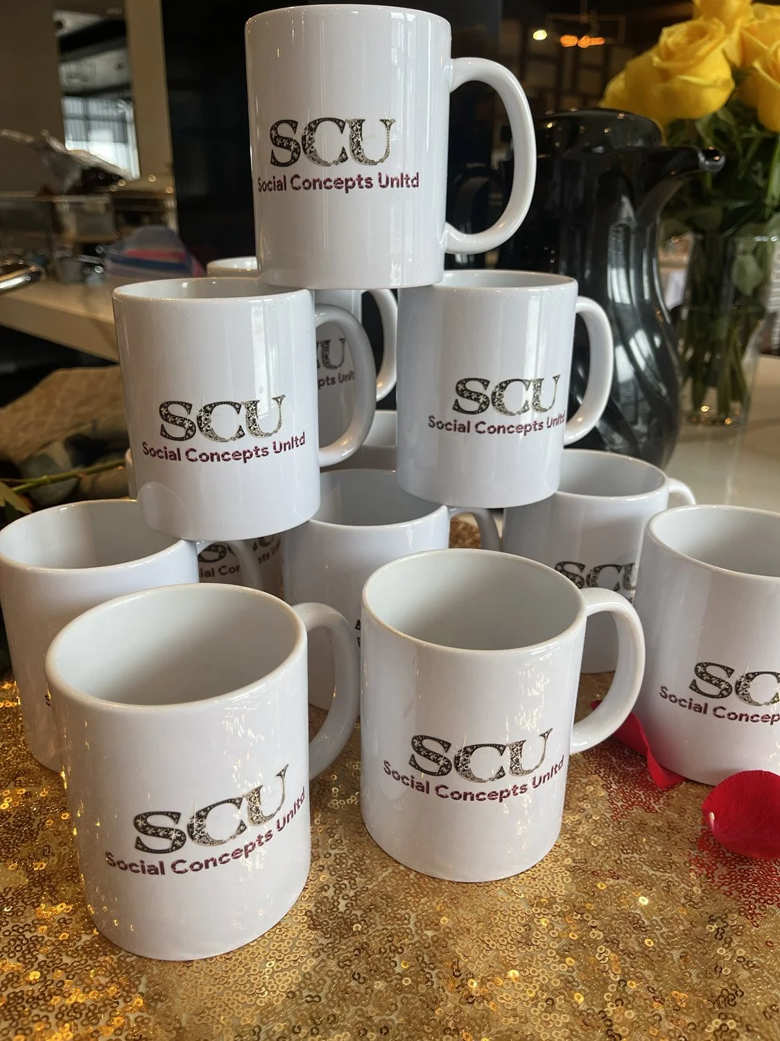 A pyramid of white coffee mugs with the logo and text 'SCU Social Concepts United' printed on them, arranged on a gold sparkly surface with a dark background including a vase of yellow roses and a black coffee pot.
