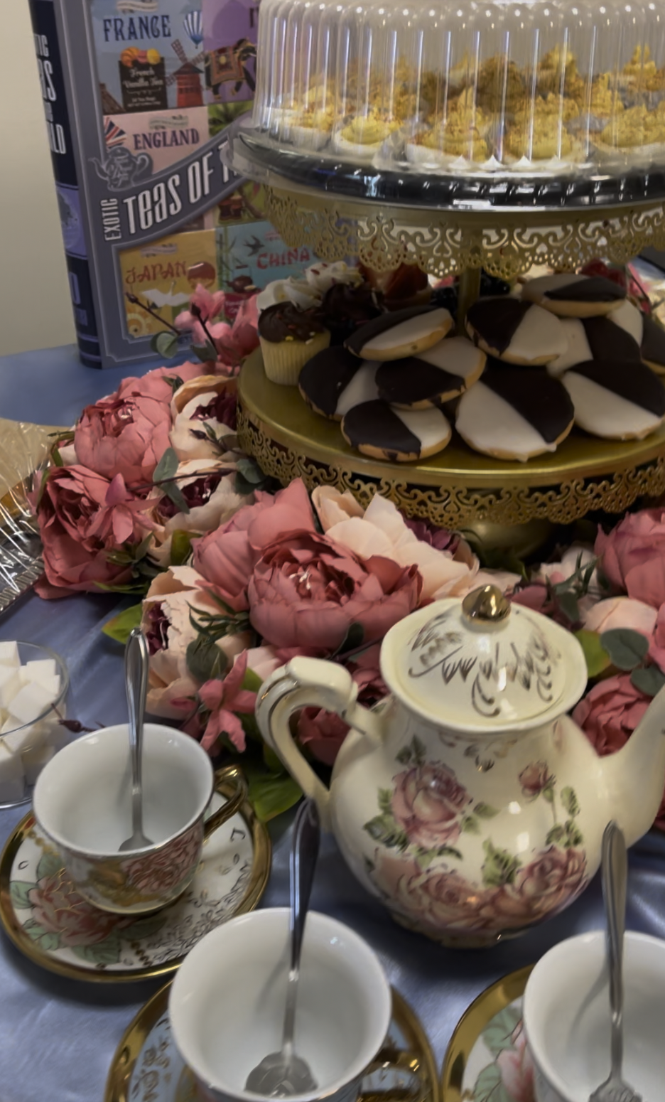 A tiered dessert tray with cookies and cupcakes, a floral teapot, teacups, and pink flowers on a table.